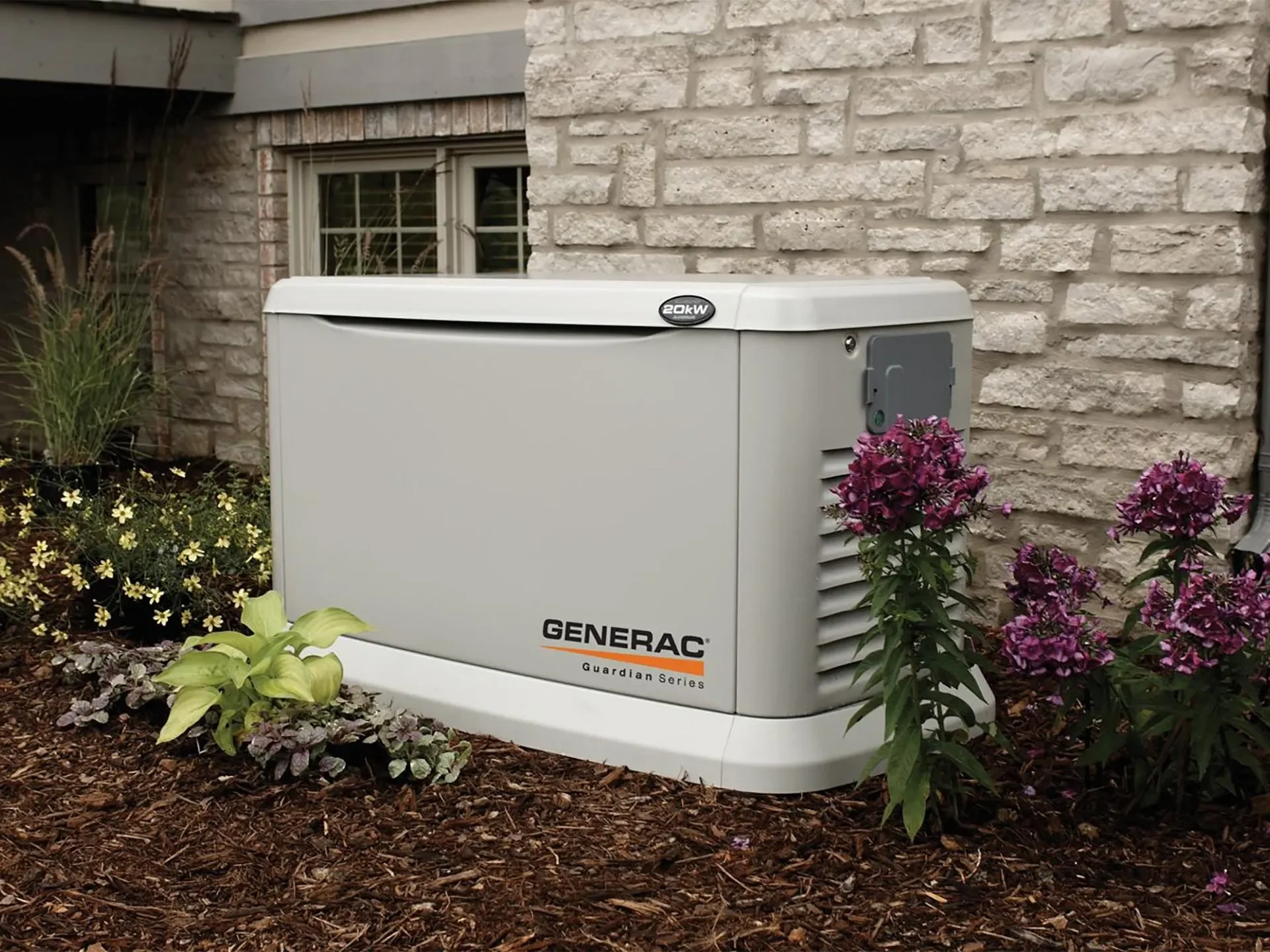 A Generac Guardian Series backup generator outside a house with a brick wall, surrounded by flowers and landscaped garden.