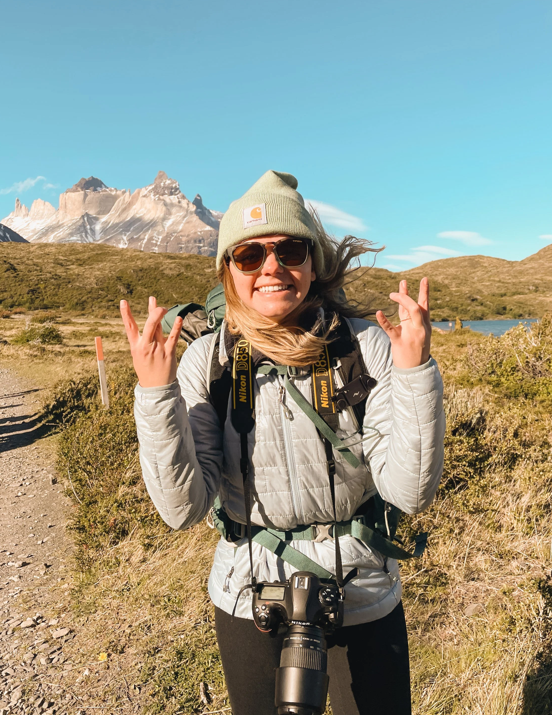 Dressed in a blue Patagonia puffy & green beanie, Anna throws up the W hand sign after finishing the ~50 mile W Trek in Patagonia's Torres del Paine National Park. With windswept hair, she grins while the mountain peaks tower behind her.