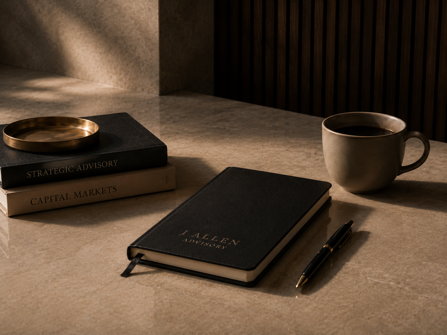 Desk with a cup of coffee, a closed black J. Allen notebook, a gold pen, and two business strategy books titled 'Strategic Advisory' and 'Capital Markets' placed on a marble surface with warm lighting.