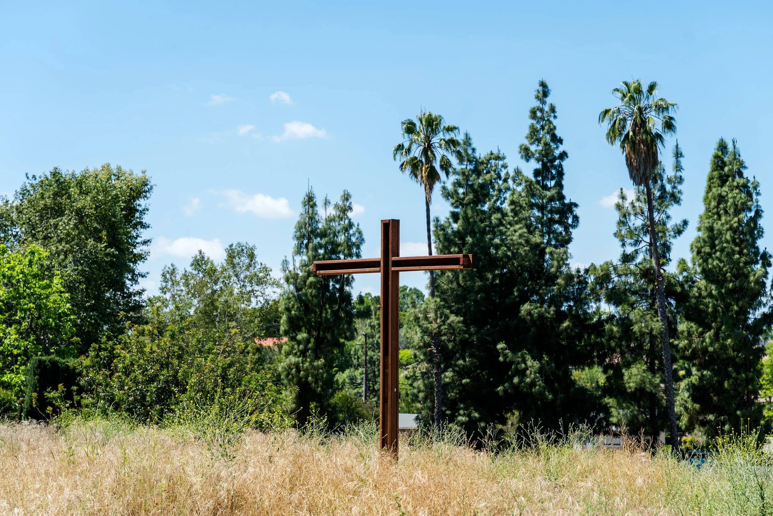 A wooden cross stands in a grassy area with various trees, including palm trees and pine trees, under a blue sky with some white clouds.