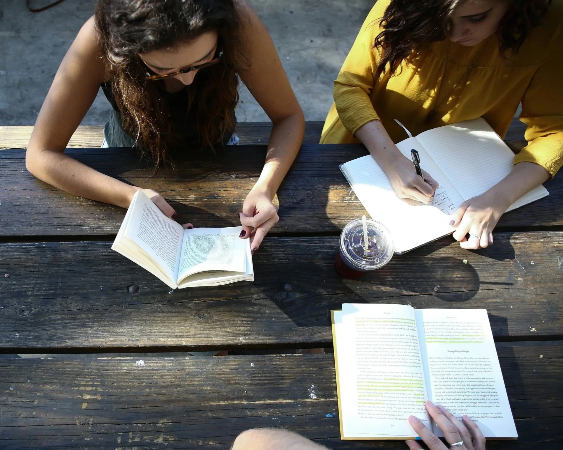 Two women sit at a wooden picnic table, reading and taking notes from their notebooks and a book, while enjoying drinks with straws. One woman wears a yellow blouse, the other a dark top, both with long, curly hair.