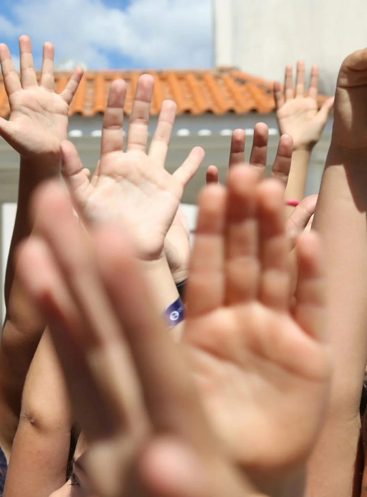 Multiple children raising their hands outdoors with a building and blue sky in the background.