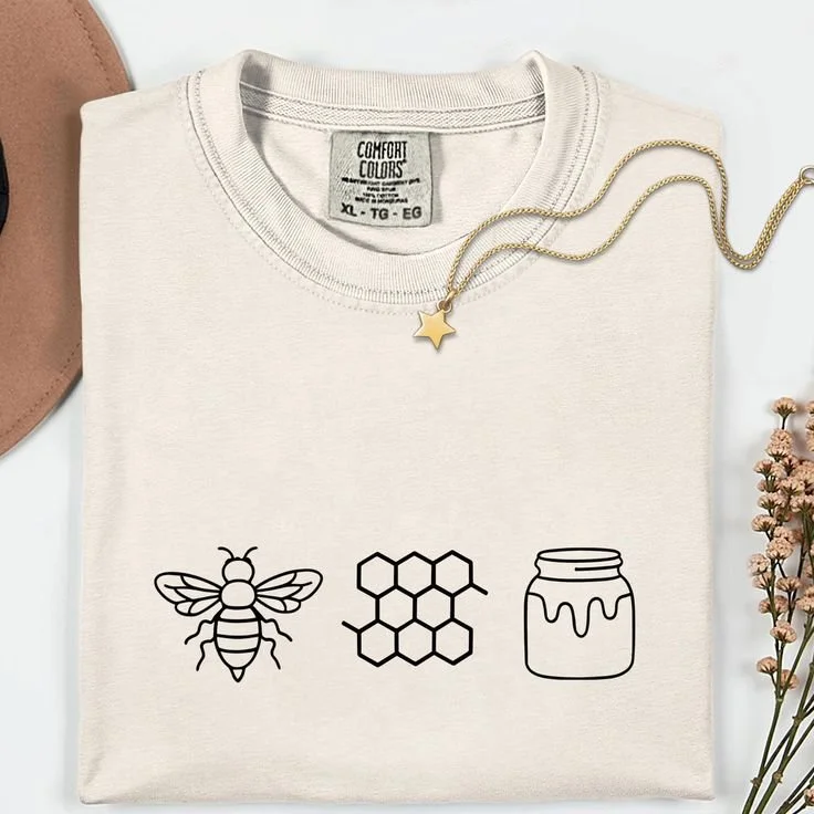 A white T-shirt with black line art of a bee, a honeycomb pattern, and a jar of honey printed on it, surrounded by a gold star necklace and some small flowers.