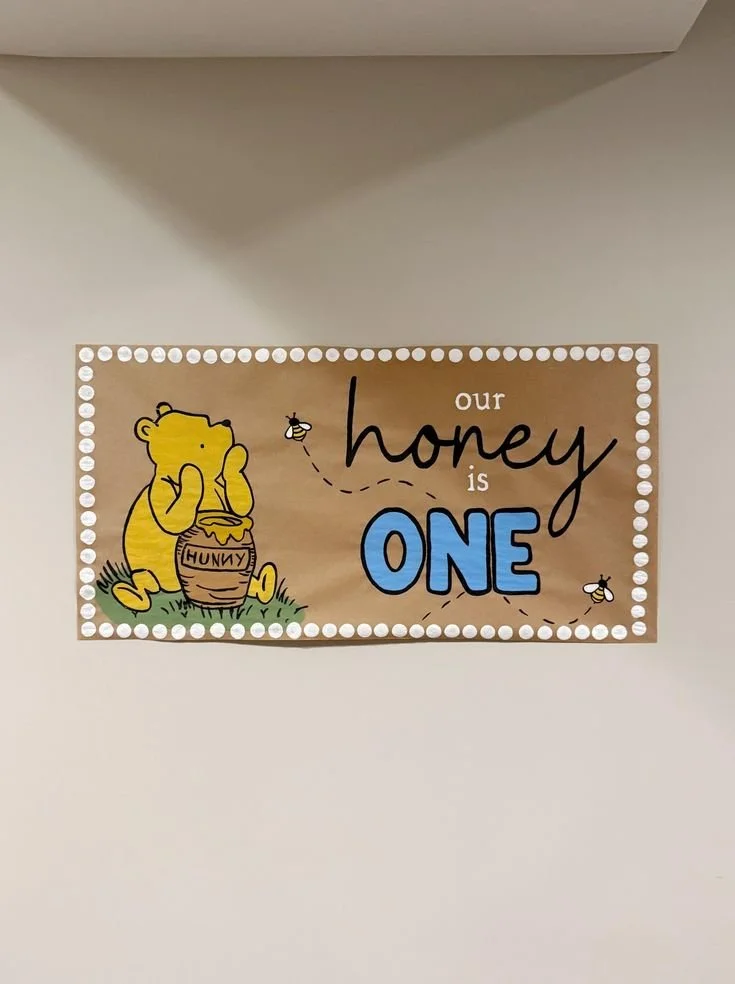 A decorative sign on a wall featuring Winnie the Pooh holding a honey jar labeled 'HUNNY,' with bees flying around, and the text 'our honey is ONE' in playful fonts.
