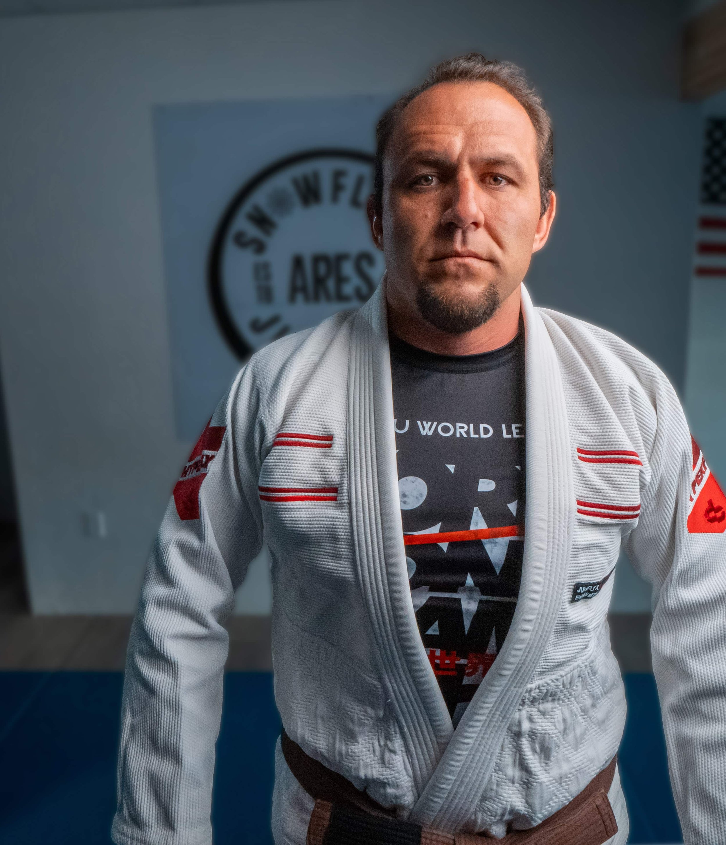 A man with short hair and a beard wearing a white martial arts gi with a patch on the shoulder, standing in front of a blue wall with a logo.