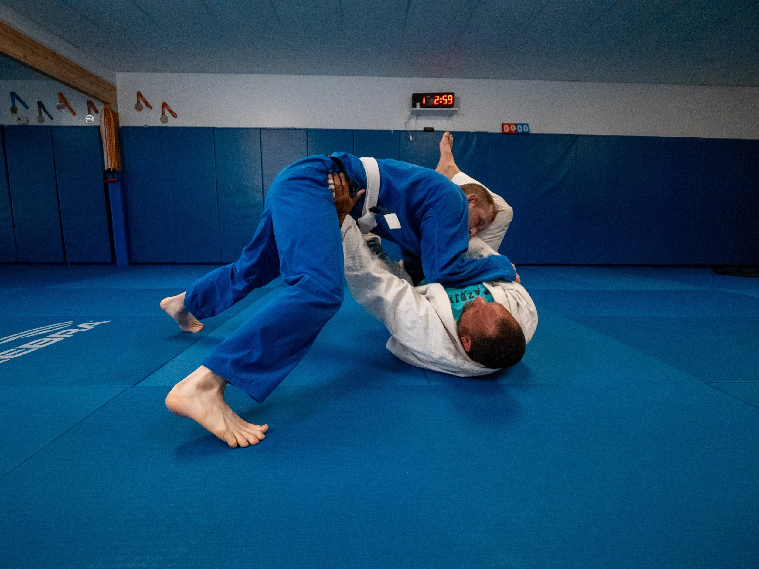 Two men practicing Brazilian Jiu-Jitsu on a blue mat in a martial arts gym, with one in a white gi and the other in a blue gi, in a grappling position.
