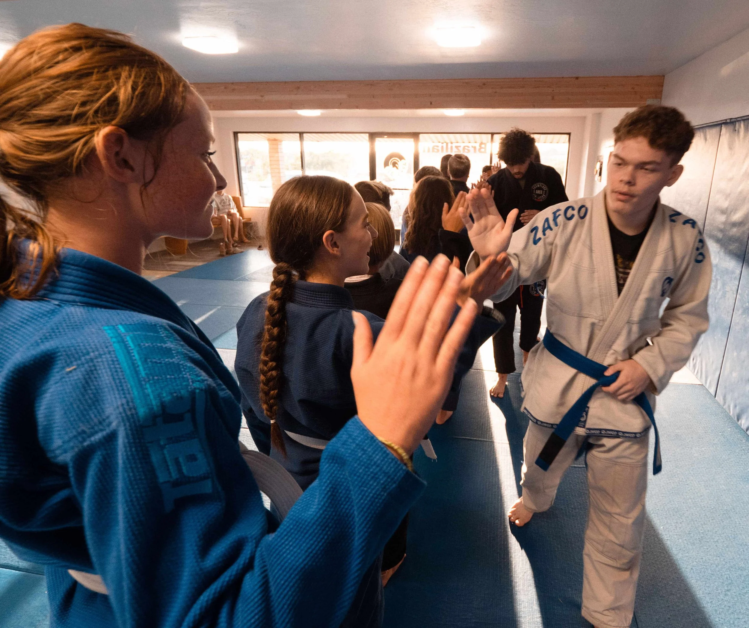 Group of martial arts students in a dojo. Two girls in blue gi practicing high-fives, a boy in a white gi with a blue belt observing, and other students in the background.