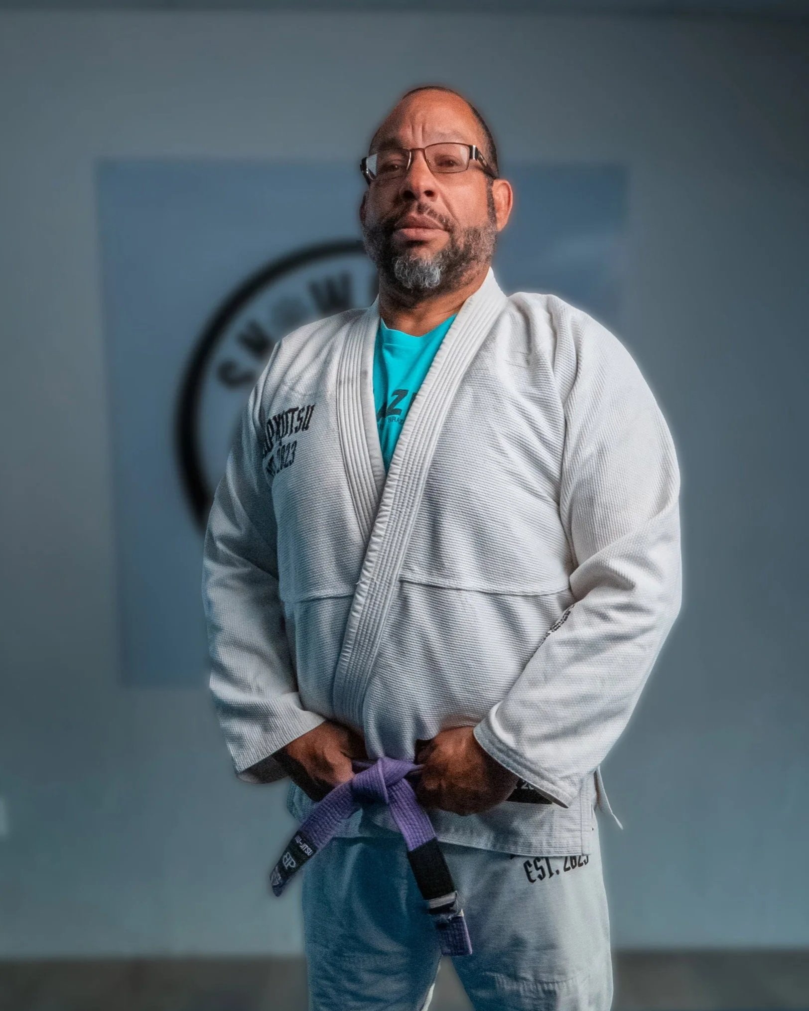 Man dressed in a white martial arts gi holding a purple belt, standing in front of a gray background with a partially visible logo.