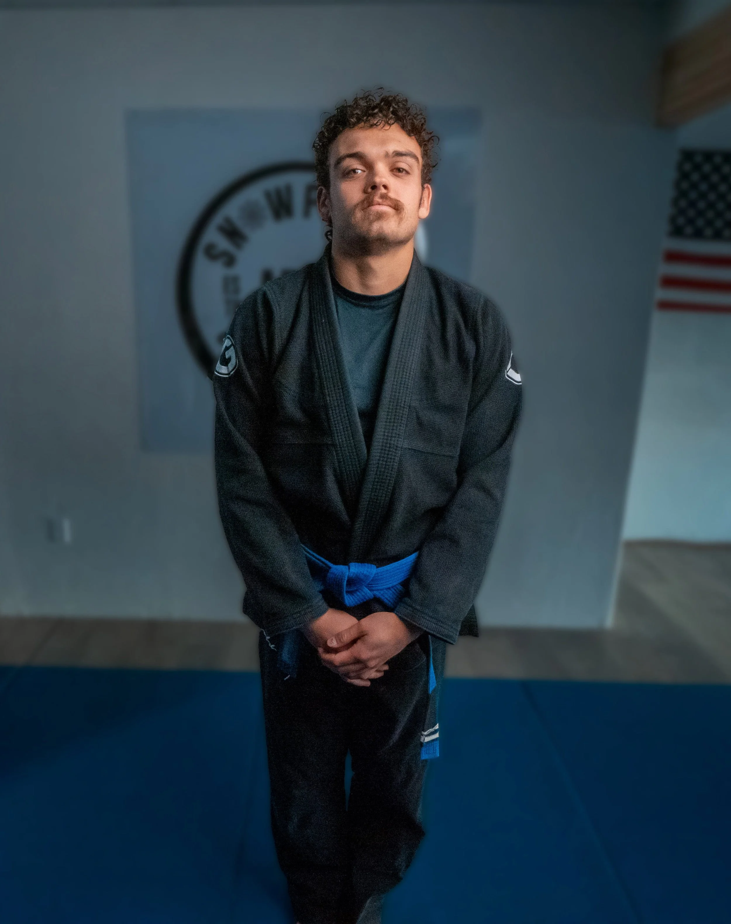 A young man with curly hair and a mustache dressed in a black martial arts gi with a blue belt, standing with his hands clasped in front of him, in a martial arts dojo with a white wall, a martial arts emblem, and an American flag in the background.