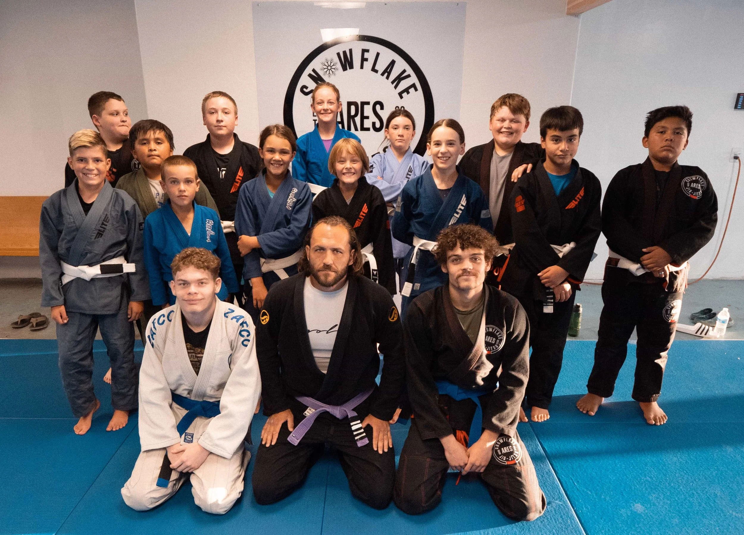 Group of children and instructors in Brazilian Jiu-Jitsu uniforms on a blue mat in a training facility, smiling for the photo.