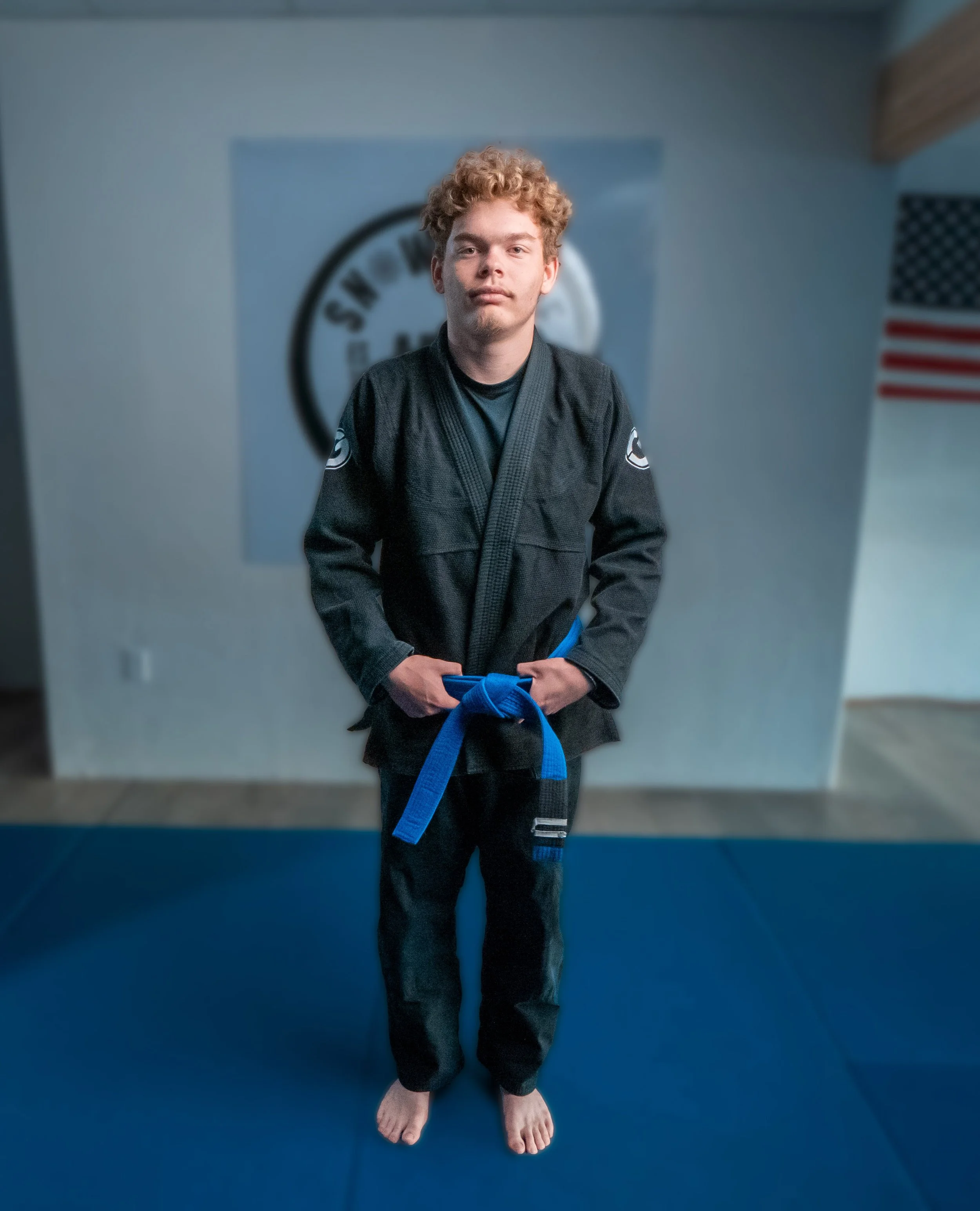 A person with curly red hair standing on a blue martial arts mat, wearing a black gi with patches, and holding a blue belt, with an American flag and martial arts logo in the background.