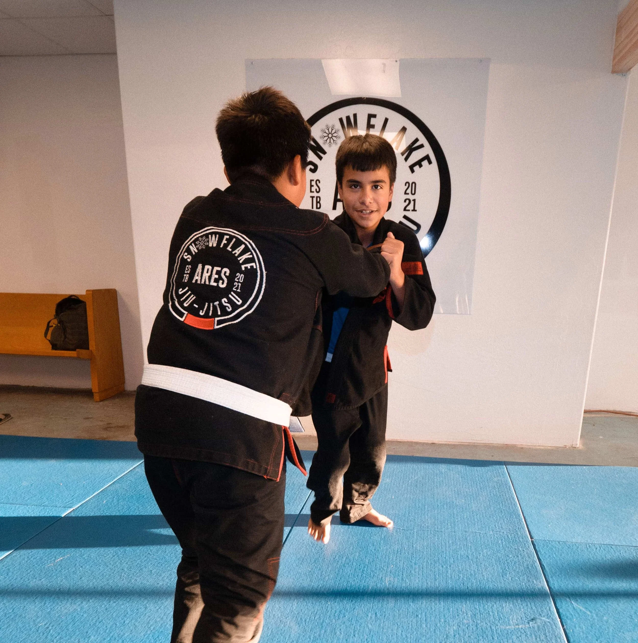 Two young boys practicing Brazilian Jiu-Jitsu in a dojo, wearing black gis with patches, on blue mats, with a white wall and a circular logo in the background.
