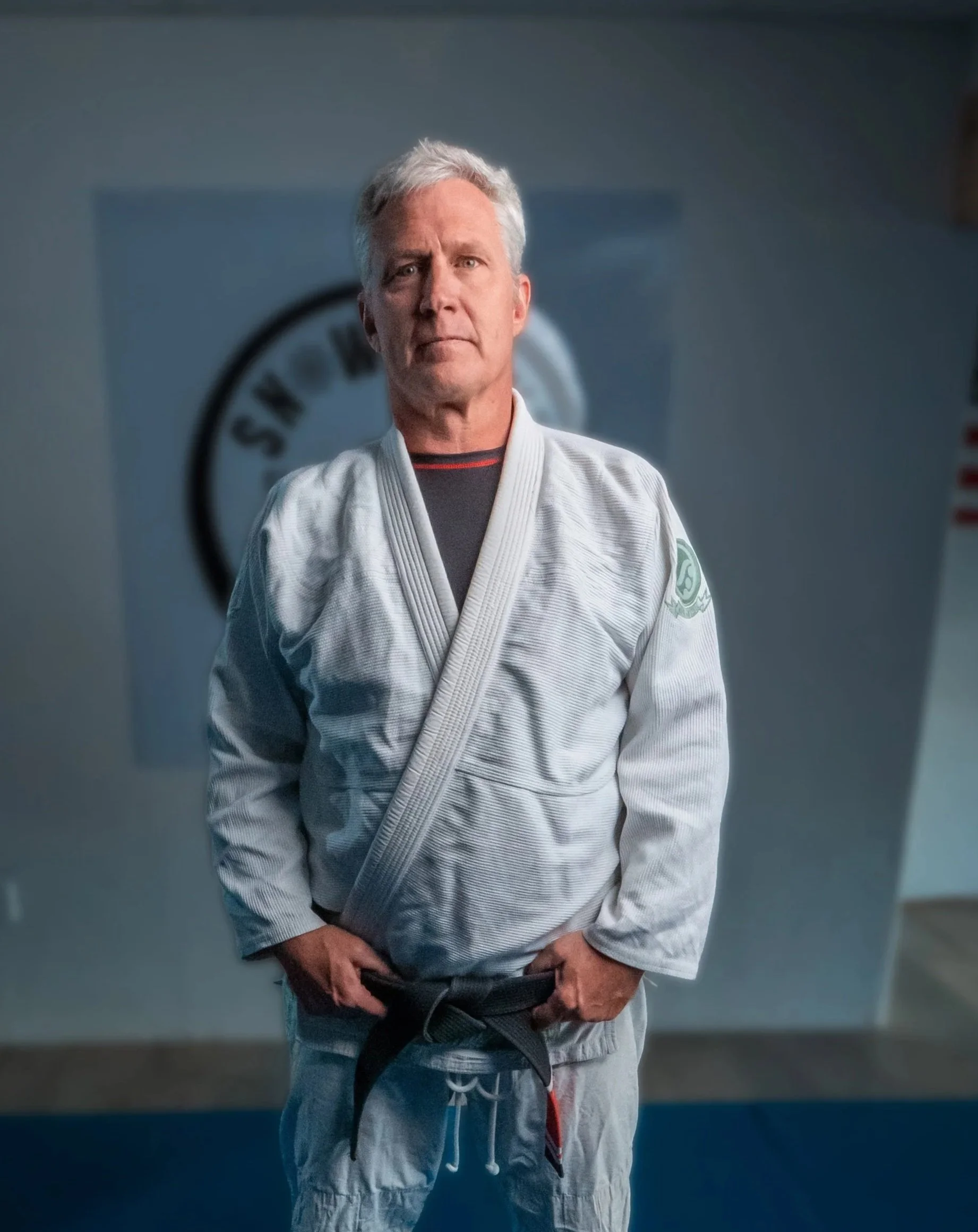 A middle-aged man with gray hair standing in a martial arts dojo, wearing a white gi and a black belt, with a serious expression.