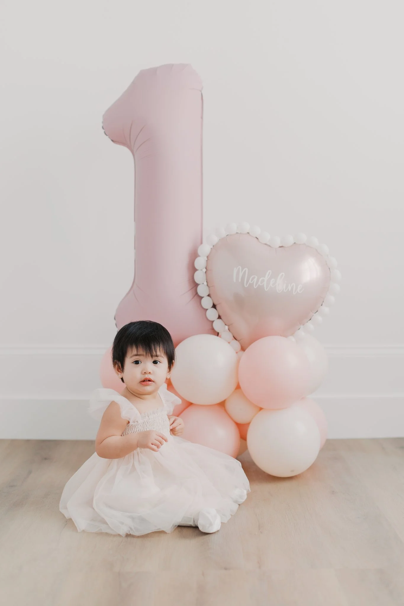 A young girl with short black hair and light skin wearing a white dress with ruffles, sitting on a light-colored wooden floor. Behind her is a decorative arrangement of pink and white balloons, including a large pink balloon shaped like the number one and a heart-shaped balloon with the name 'Madeline' written on it, surrounded by smaller balloons.