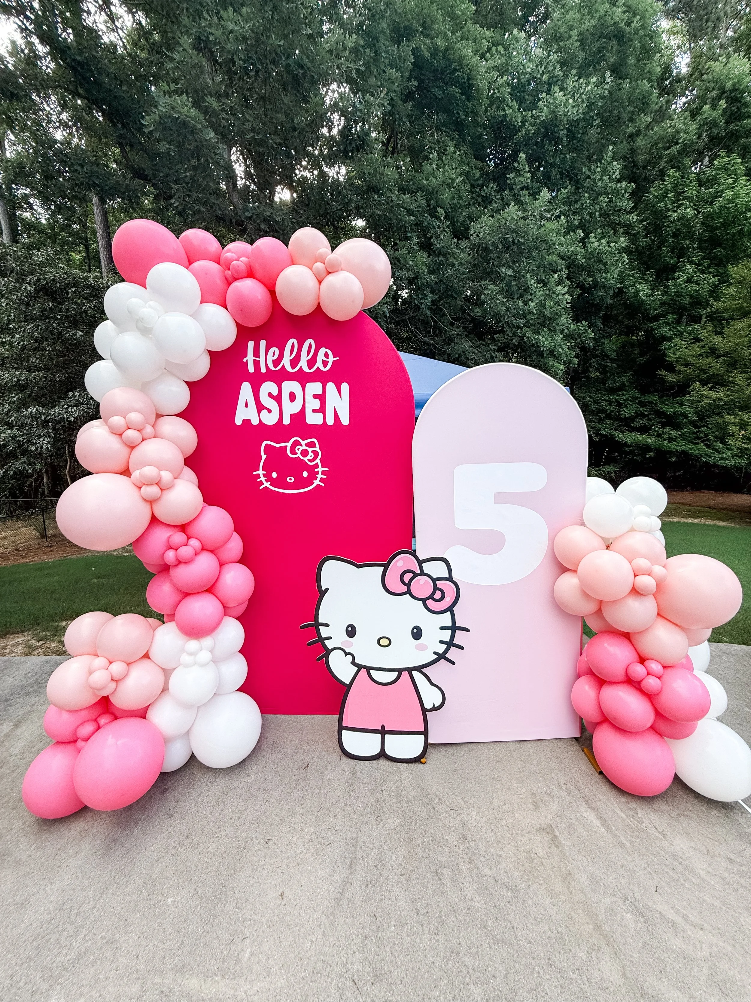 Decorative display for a Hello Kitty-themed event with a pink oval sign saying 'Hello Aspen' surrounded by pink and white balloons with cluster arrangement, a cartoon Hello Kitty figure, a large white letter 'F', and trees in the background.