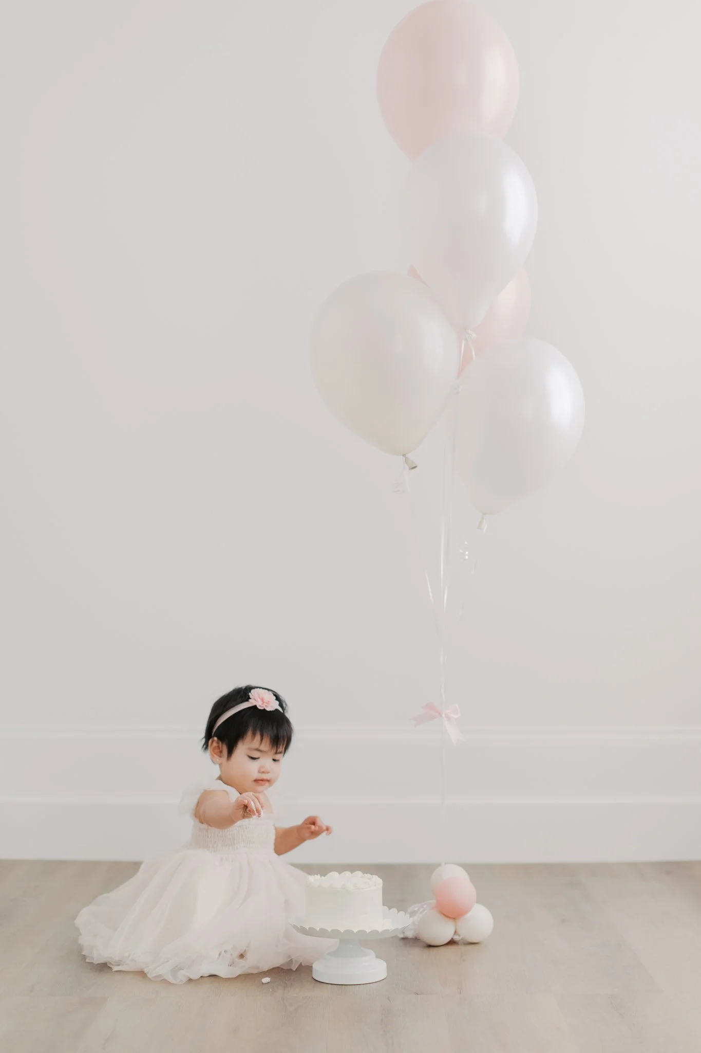 A young girl in a white dress and pink headband with a flower is sitting on the floor, surrounded by pink and white balloons and a cake, in a minimalistic room with white walls. The girl is reaching towards the cake.