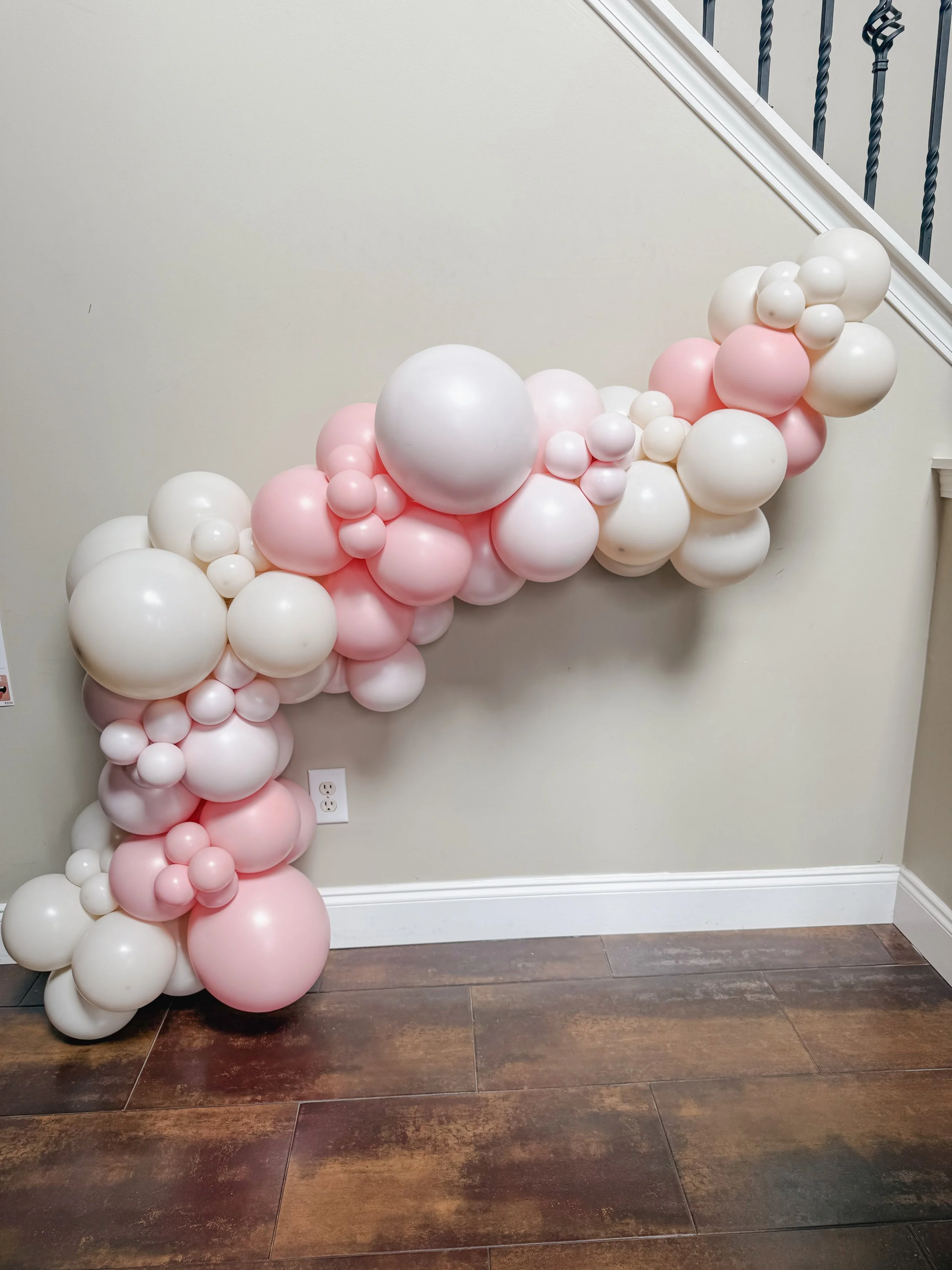 A balloon cascade with white and pink balloons, arranged on a stairway railing and wall, with a dark hardwood floor.