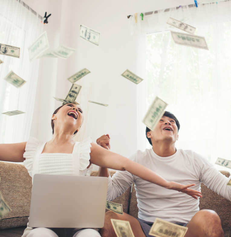 Happy couple sitting on a sofa with a laptop, throwing dollar bills in the air.