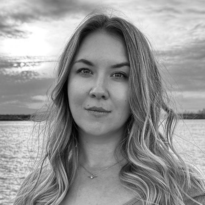 A young woman with long, wavy hair standing outdoors near water during sunset, with clouds in the sky.