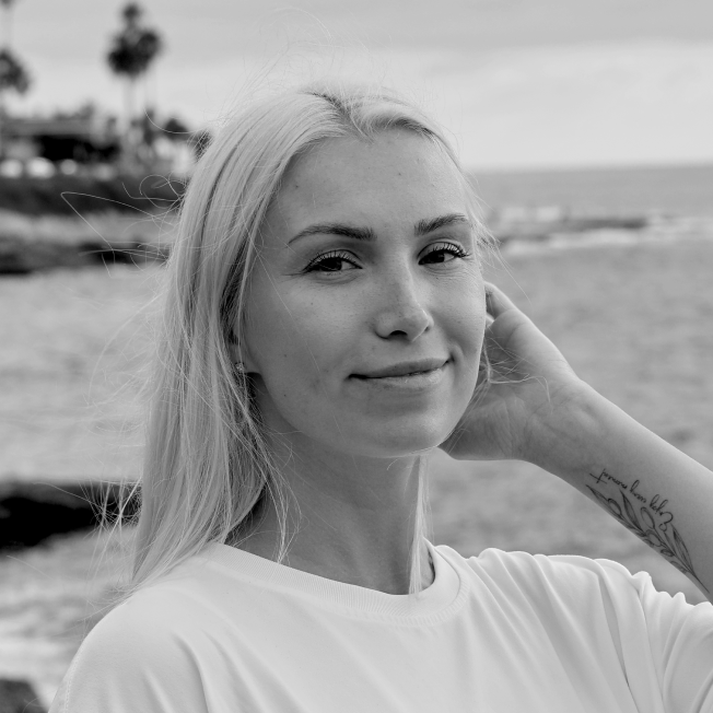 A young woman with long blonde hair smiling slightly at the camera on a beach, with ocean waves and palm trees in the background, in black and white.