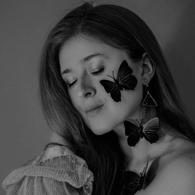 A woman with eyes closed, posing with three large butterflies on her face and neck in a black and white photo.