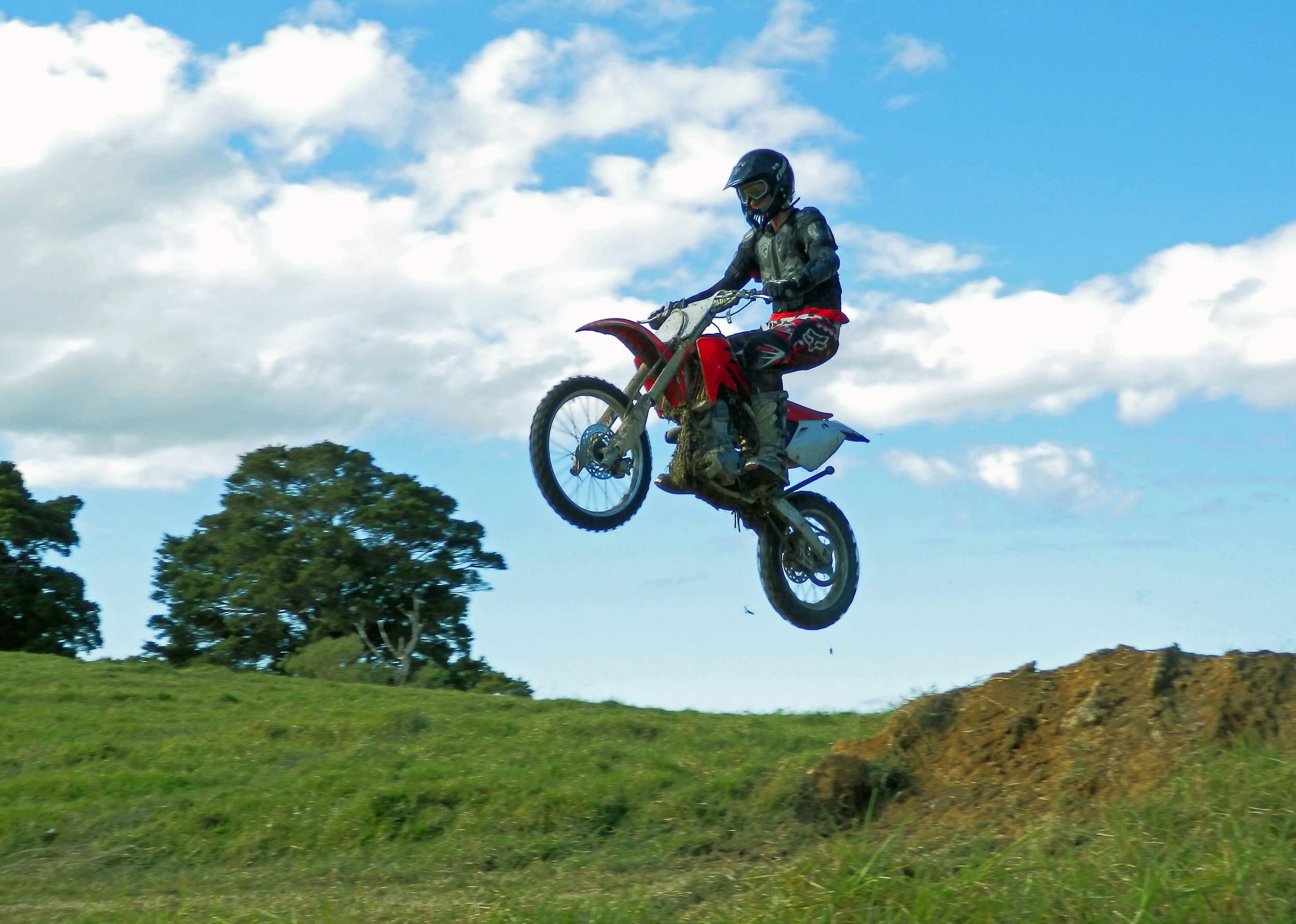 A person wearing a helmet and protective gear performing a stunt on a dirt bike in mid-air over grassy terrain under a blue sky with clouds.