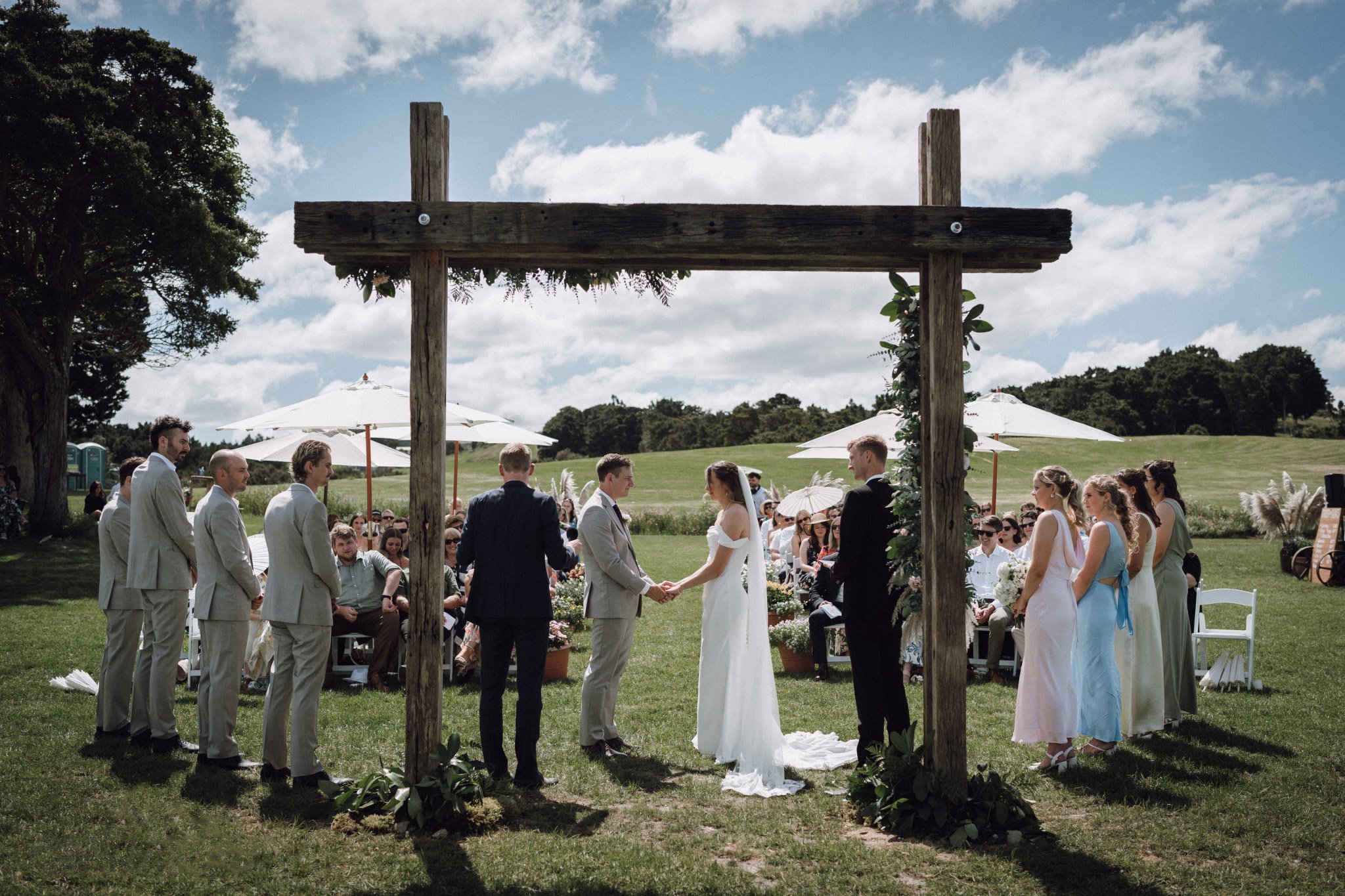 A couple getting married outdoors under a wooden arch, with guests seated under umbrellas in the background.