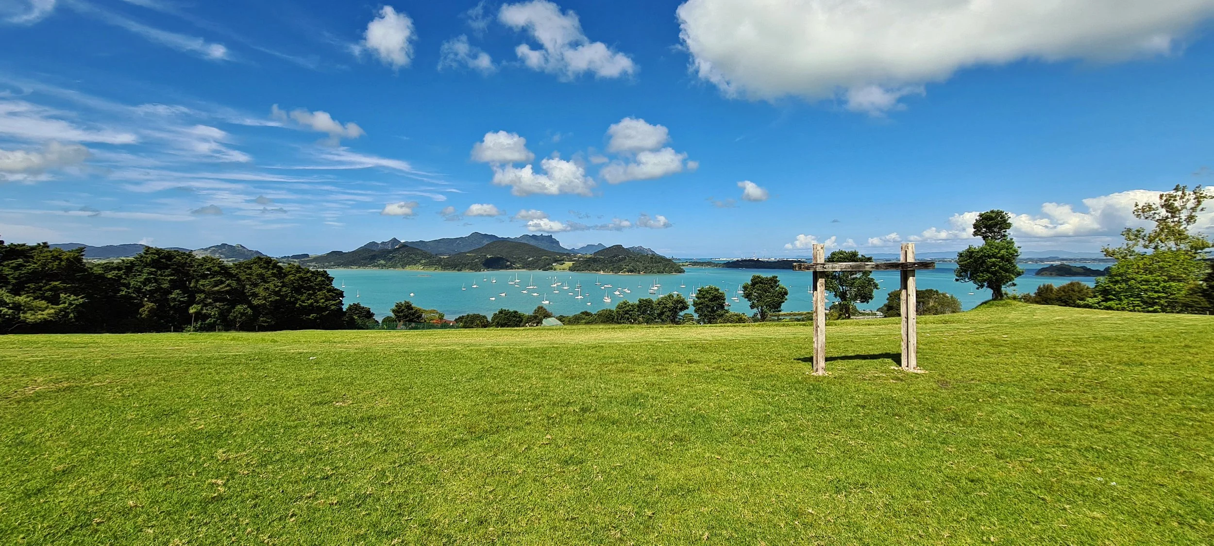 A scenic landscape featuring a lush green field with a wooden goalpost in the foreground, a dense line of trees, a large body of water with sailboats, and mountain ranges under a partly cloudy blue sky.