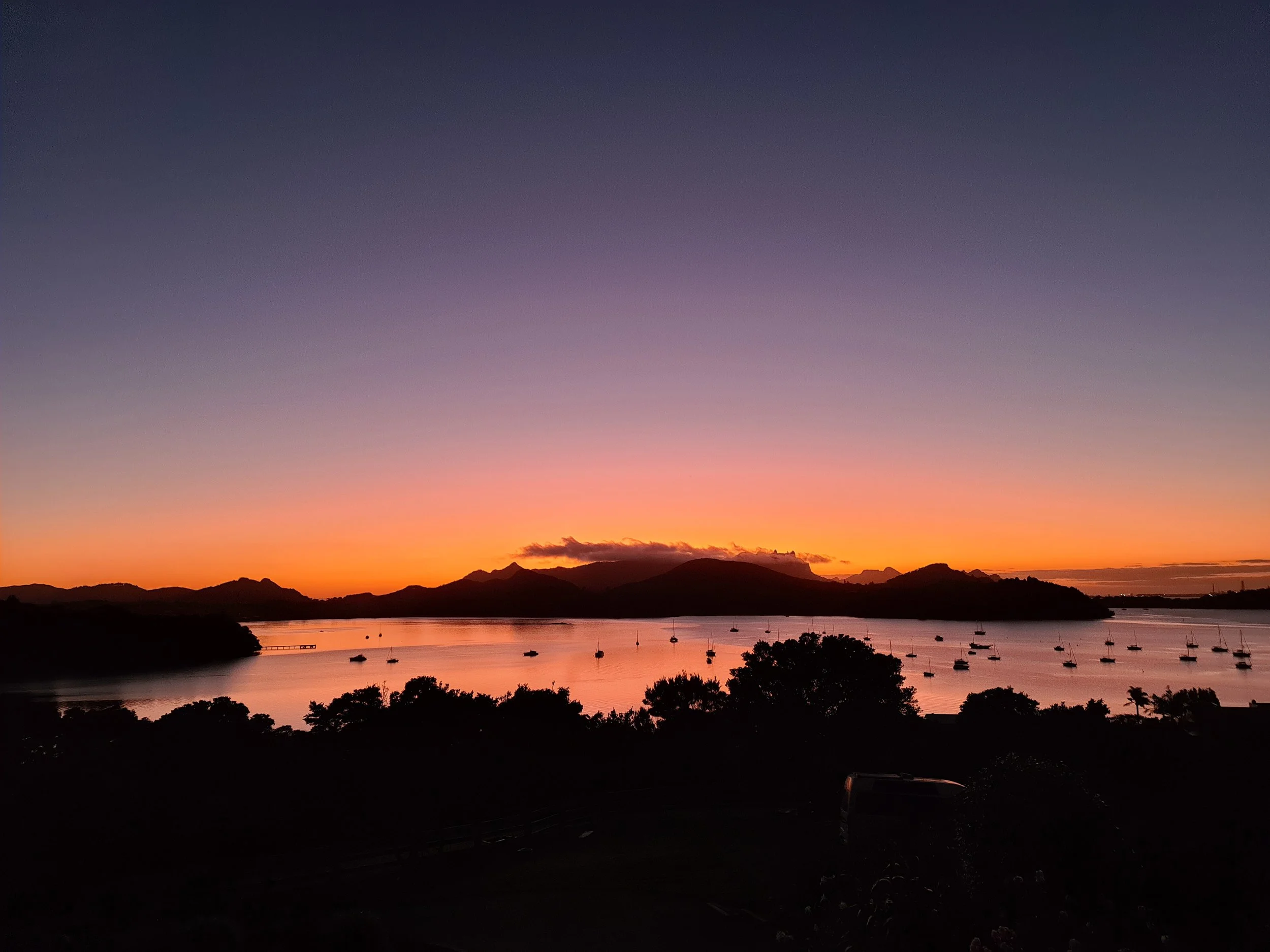 View of a serene body of water with sailboats at sunset, silhouetted hills in the background, and a colorful sky transitioning from purple to orange.