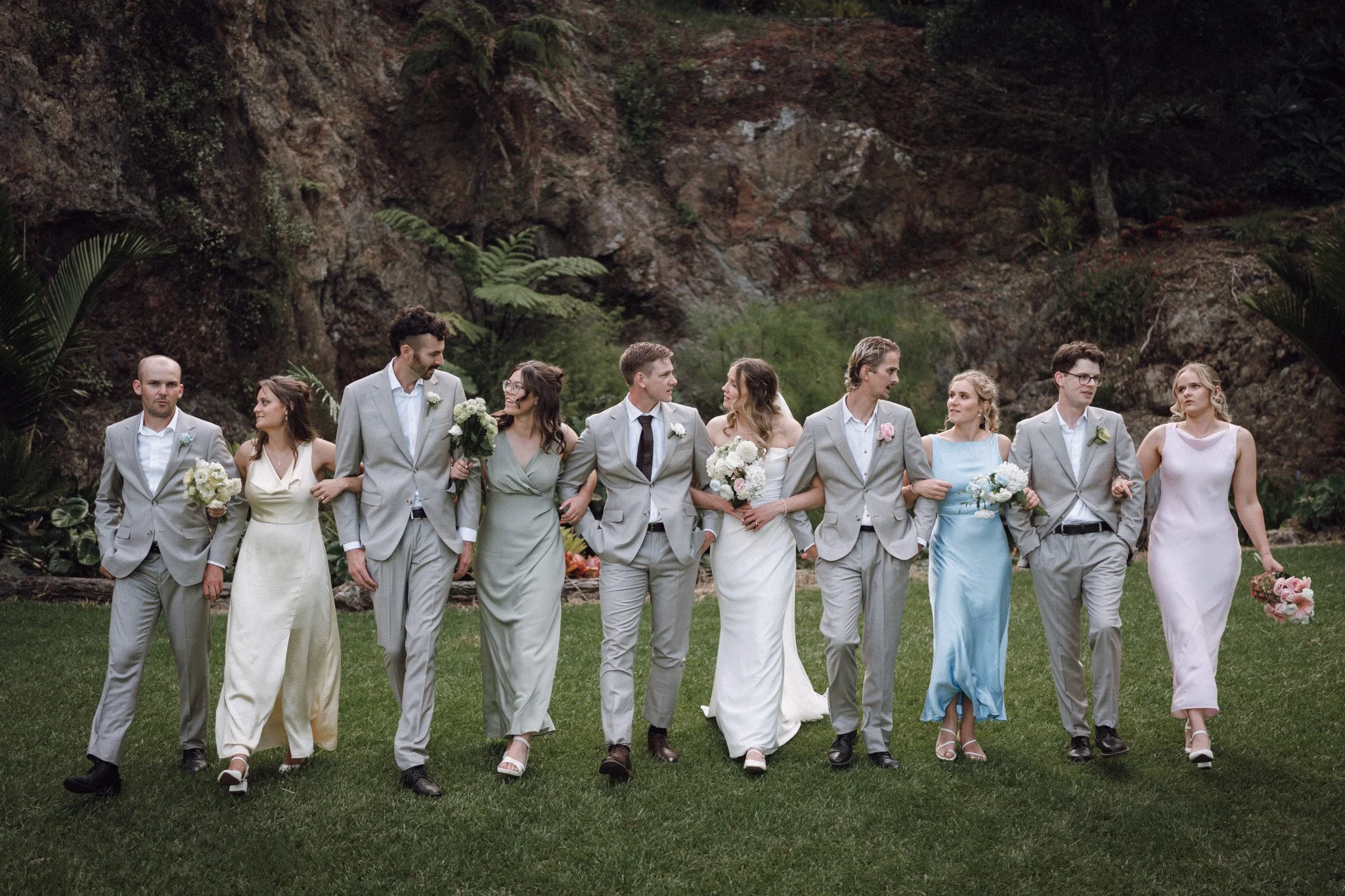 A group of ten people appears to be at a wedding, dressed in formal attire and walking arm in arm outdoors on a grassy area. The background features a rocky cliff with greenery.
