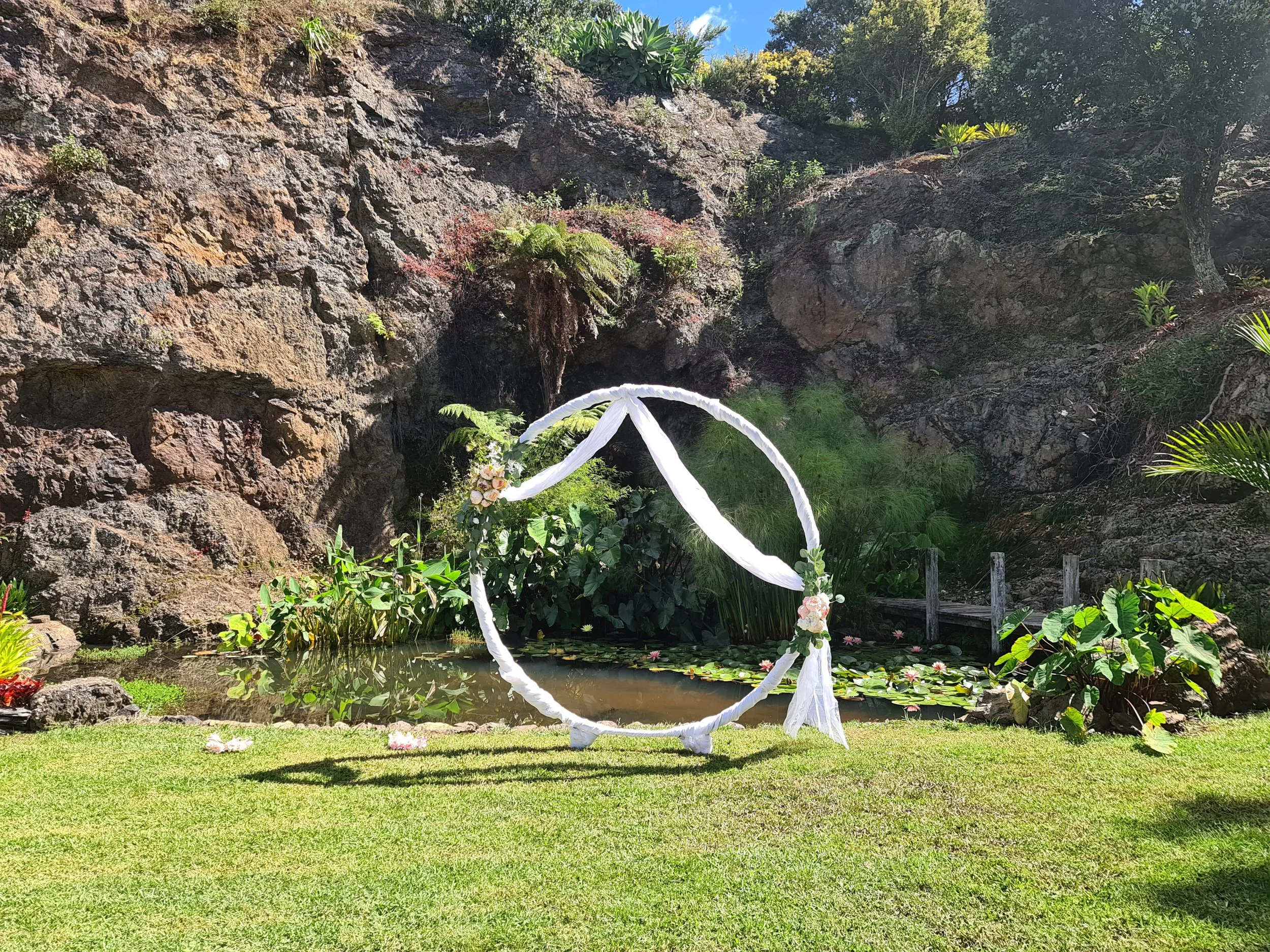 Outdoor wedding setup with a circular floral arch near a small pond, surrounded by lush greenery and rocky cliffs.