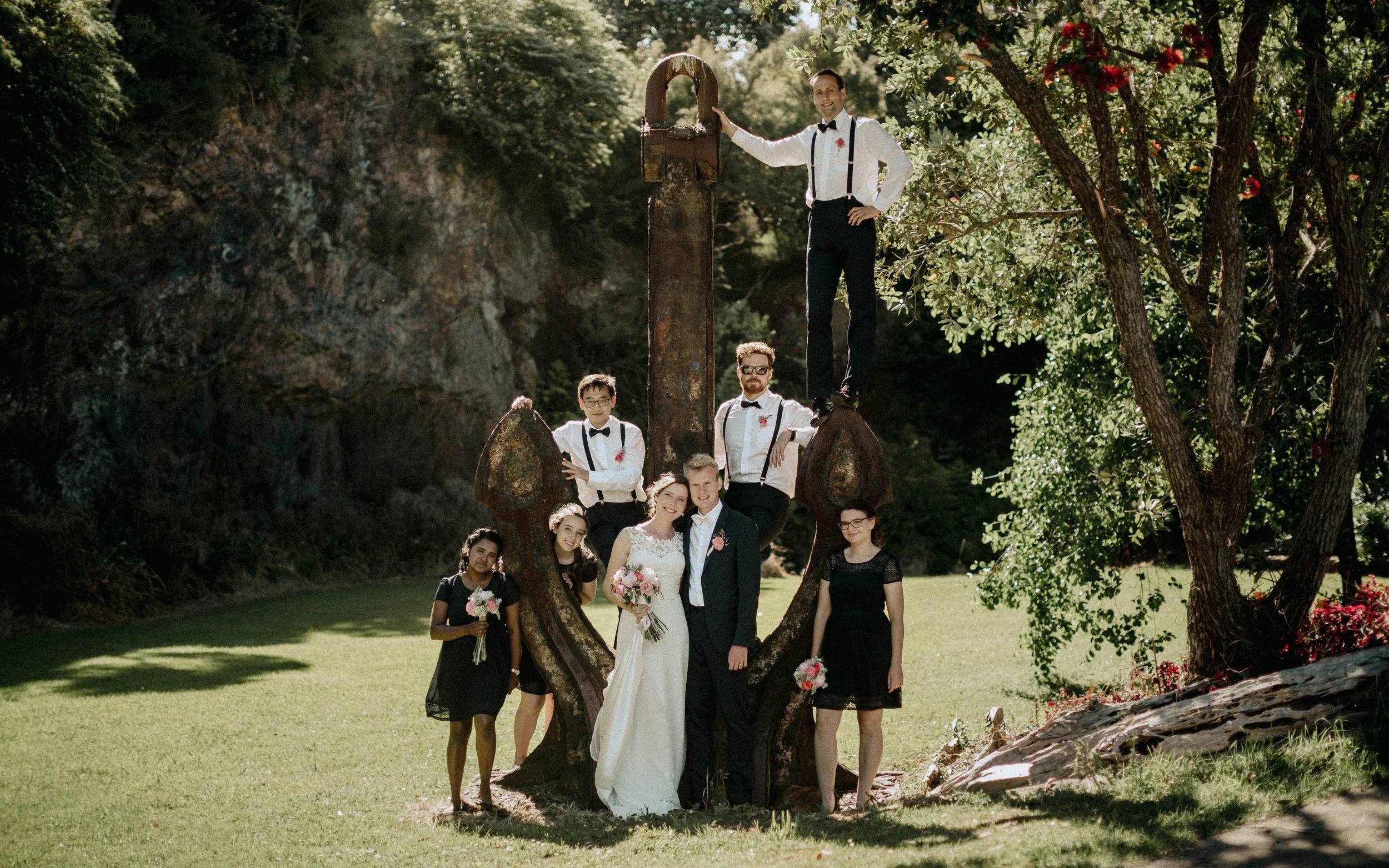Group of people gathered outdoors under a large tree during a wedding celebration, with the bride and groom in the center, surrounded by bridesmaids and groomsmen, engaged in a fun photo pose around a large, weathered wheel. The setting is sunny and green, with natural scenery in the background.