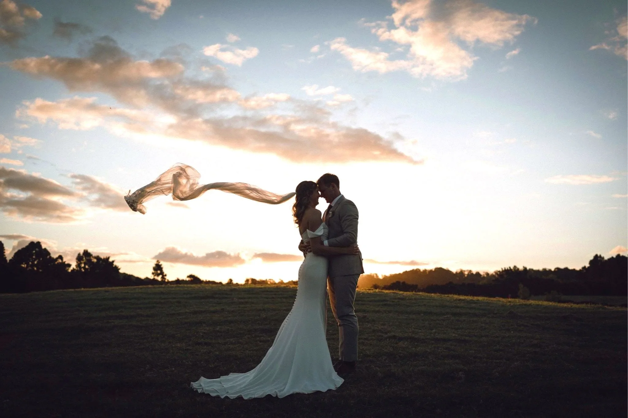 A couple embracing outdoors during sunset, with a woman in a white gown and a man in a suit. The woman's veil flows in the wind, and they stand on grass with a backdrop of trees and cloudy sky.