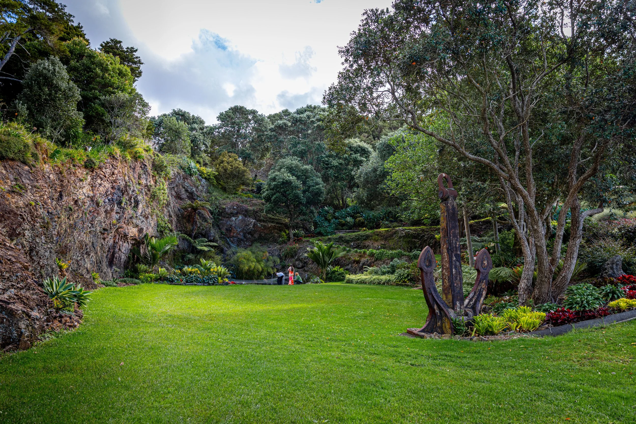 A lush, green garden with a vibrant lawn, surrounded by trees and plants, with people sitting near a pond in the background.