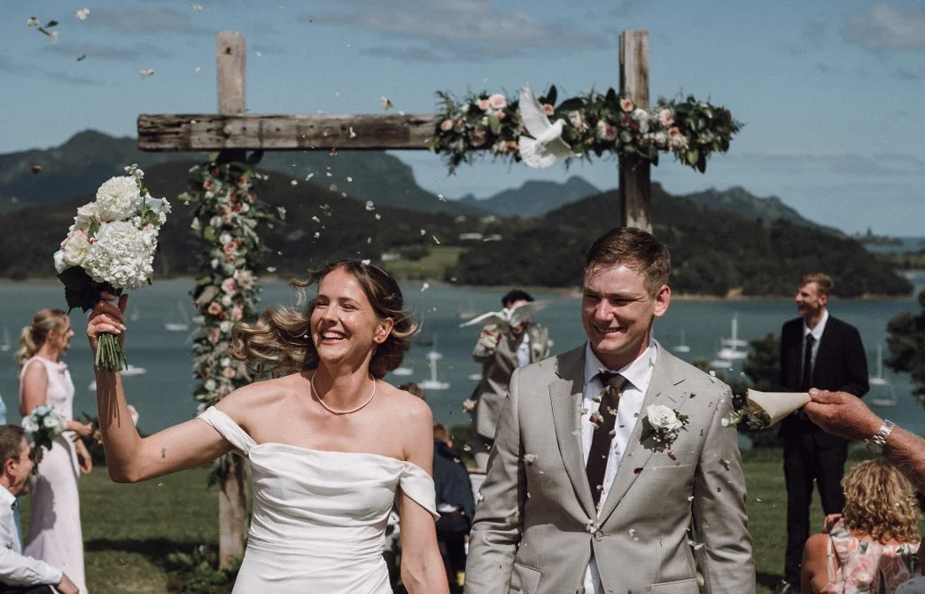 A bride and groom smiling as they walk down the aisle at an outdoor wedding ceremony by a lake with mountains in the background, under a decorated wooden arch.