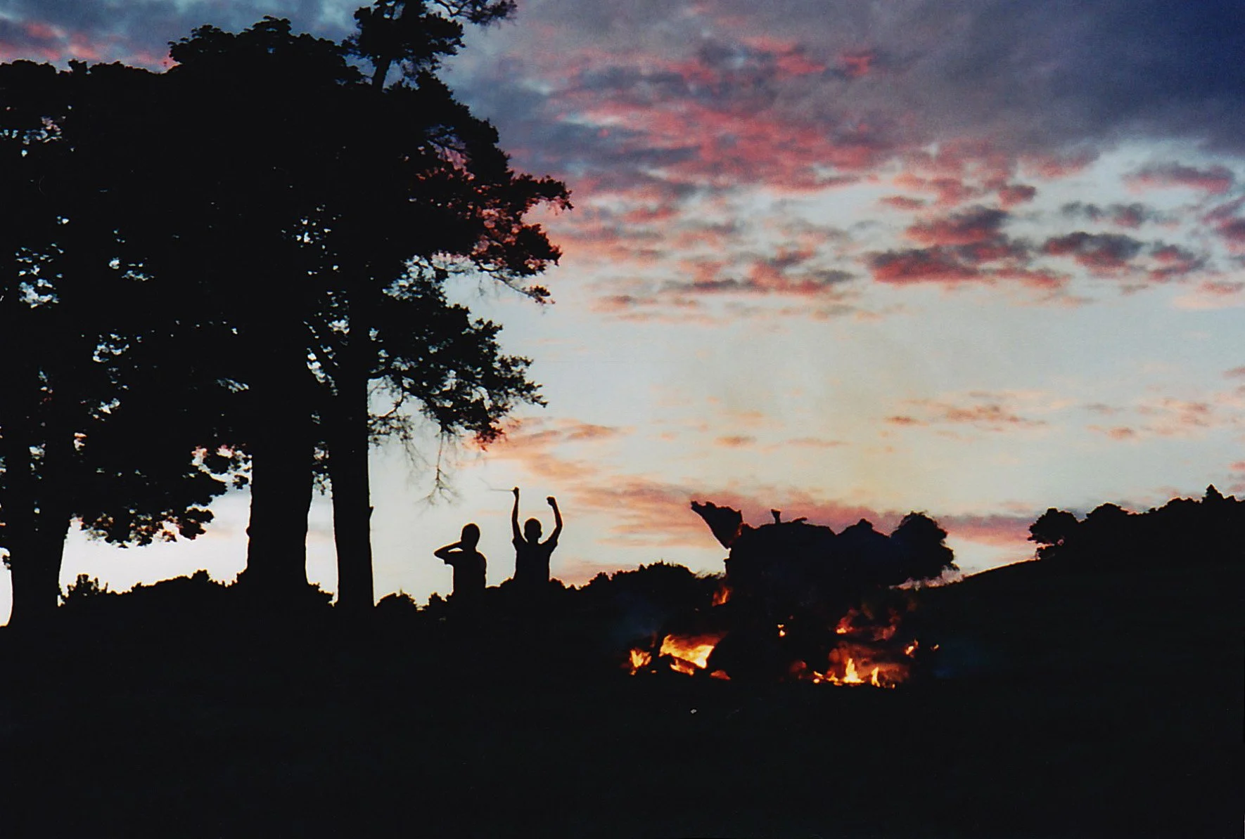 Silhouettes of three people sitting by a campfire under a colorful sunset sky with scattered clouds and trees in the background.