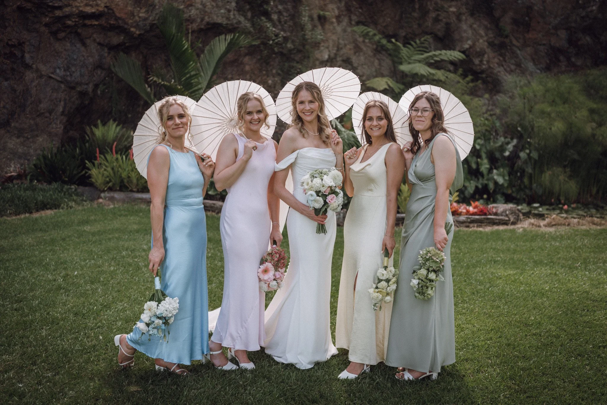 A group of five women in pastel-colored dresses standing outdoors on grass, holding white umbrellas and floral bouquets, with a natural backdrop of rocks and greenery, smiling at the camera.