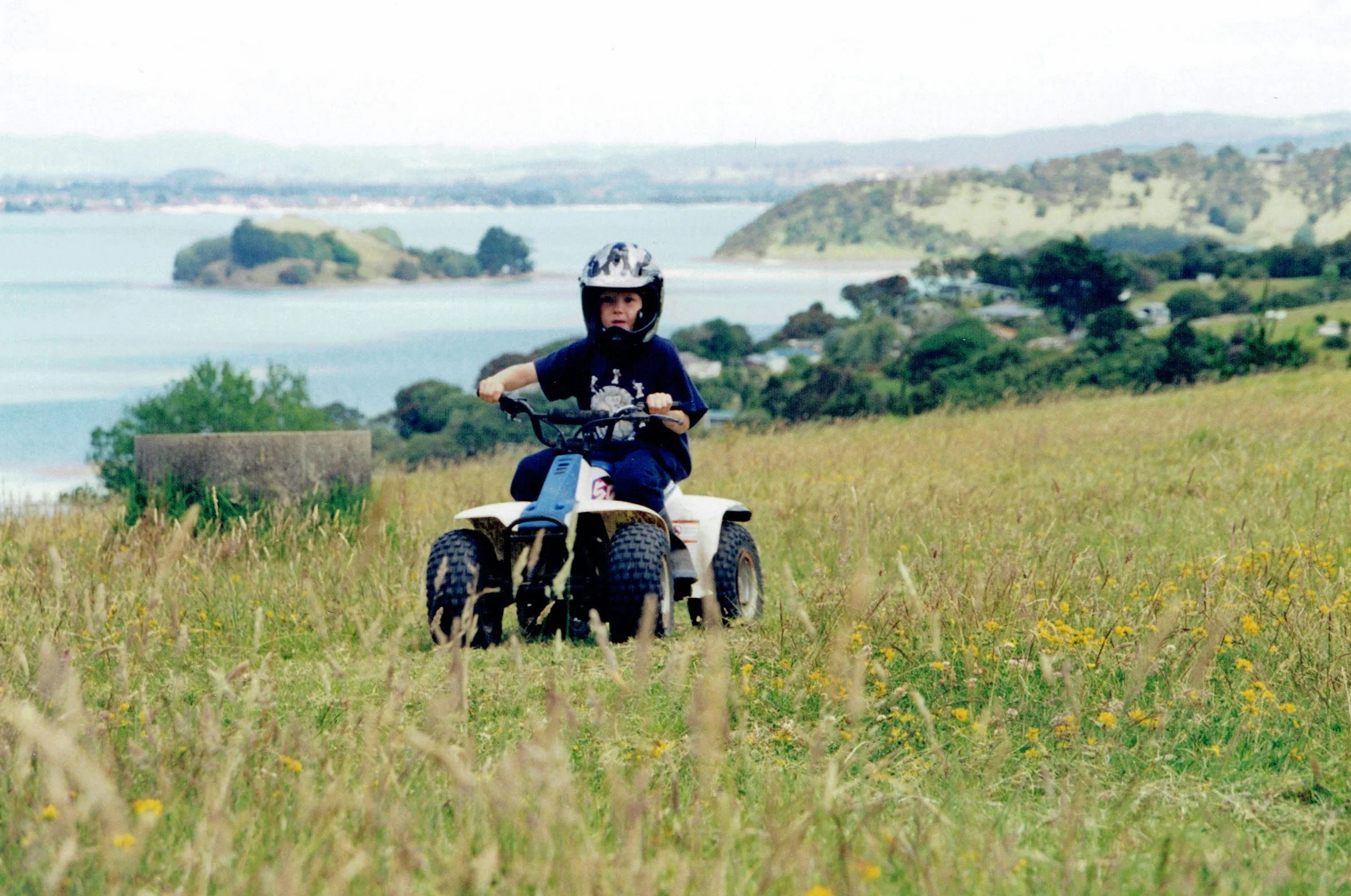 A young boy riding a quad bike through a grassy field with yellow flowers, with water, islands, and hills in the background.