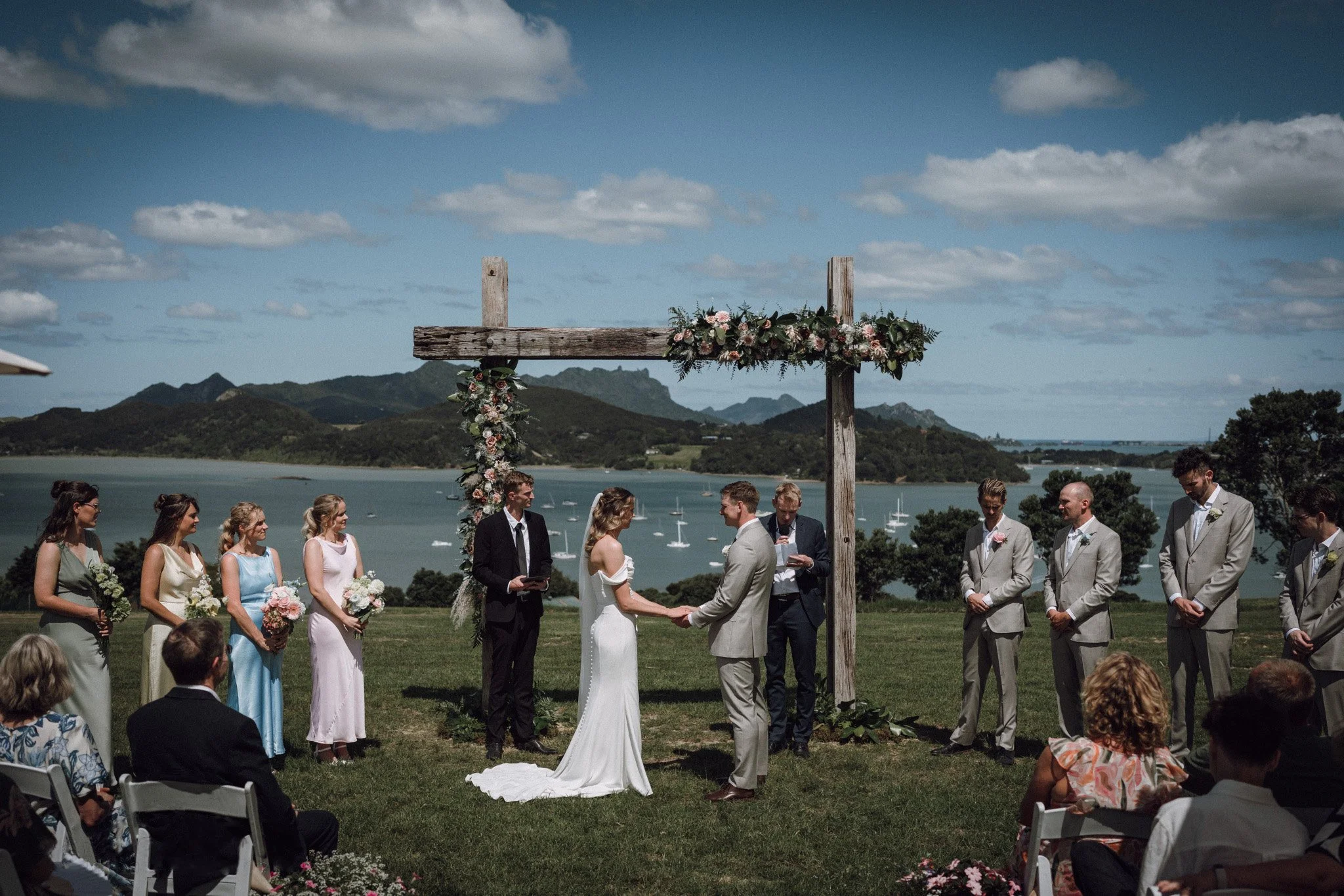 A wedding ceremony outdoors by a harbour with a mountain backdrop, showing a bride and groom holding hands under a wooden arch decorated with flowers, surrounded by wedding party and guests.