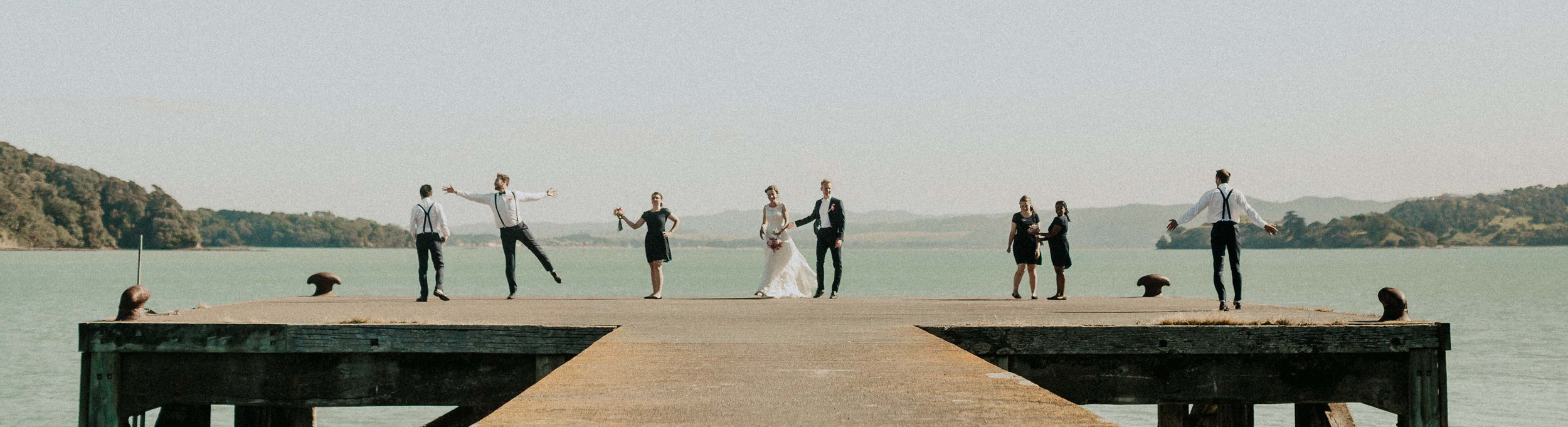 A wedding party standing on a dock over water with hills in the background.