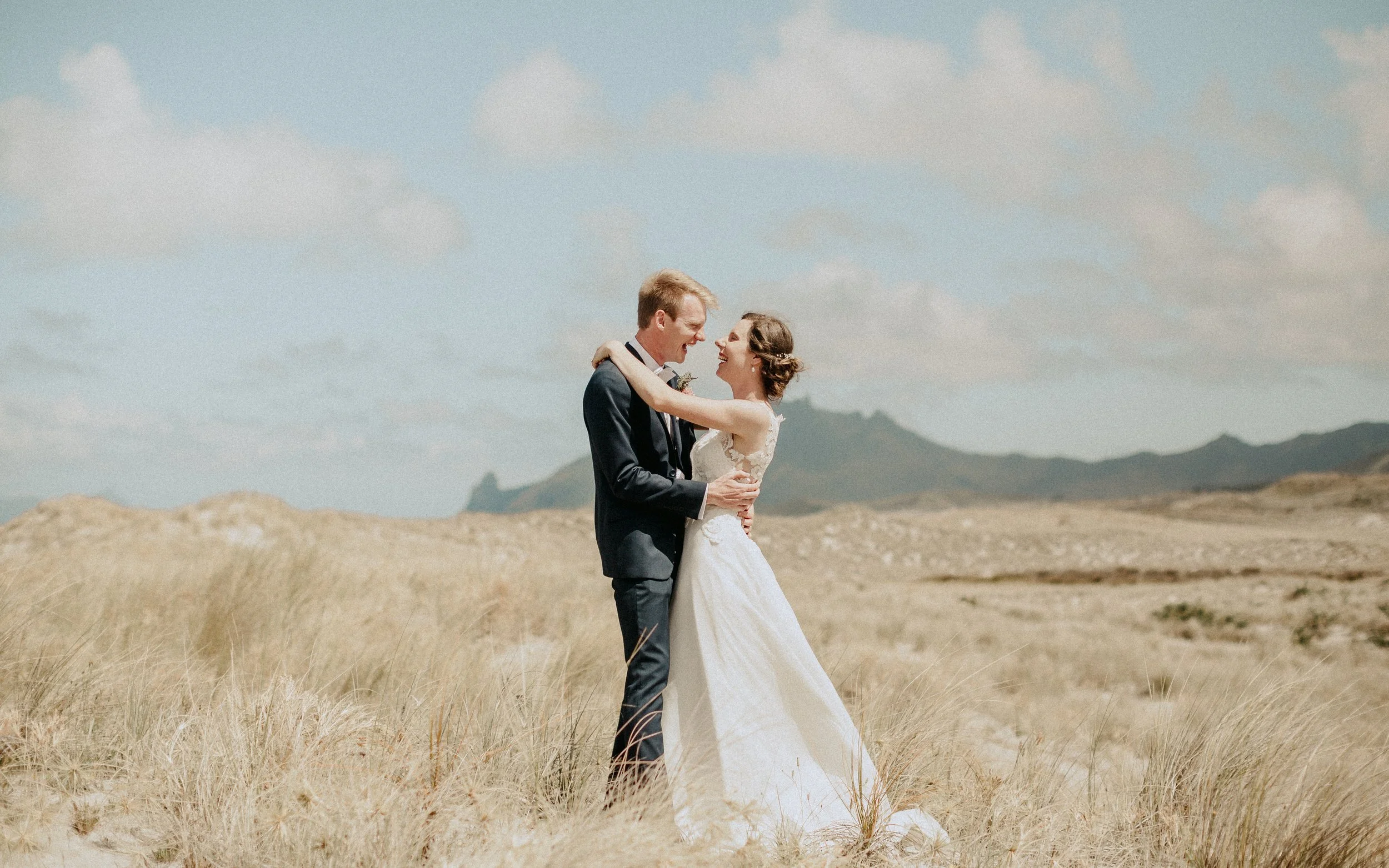 A happy couple in wedding attire embracing outdoors in a grassy field with mountains in the background under a partly cloudy sky.