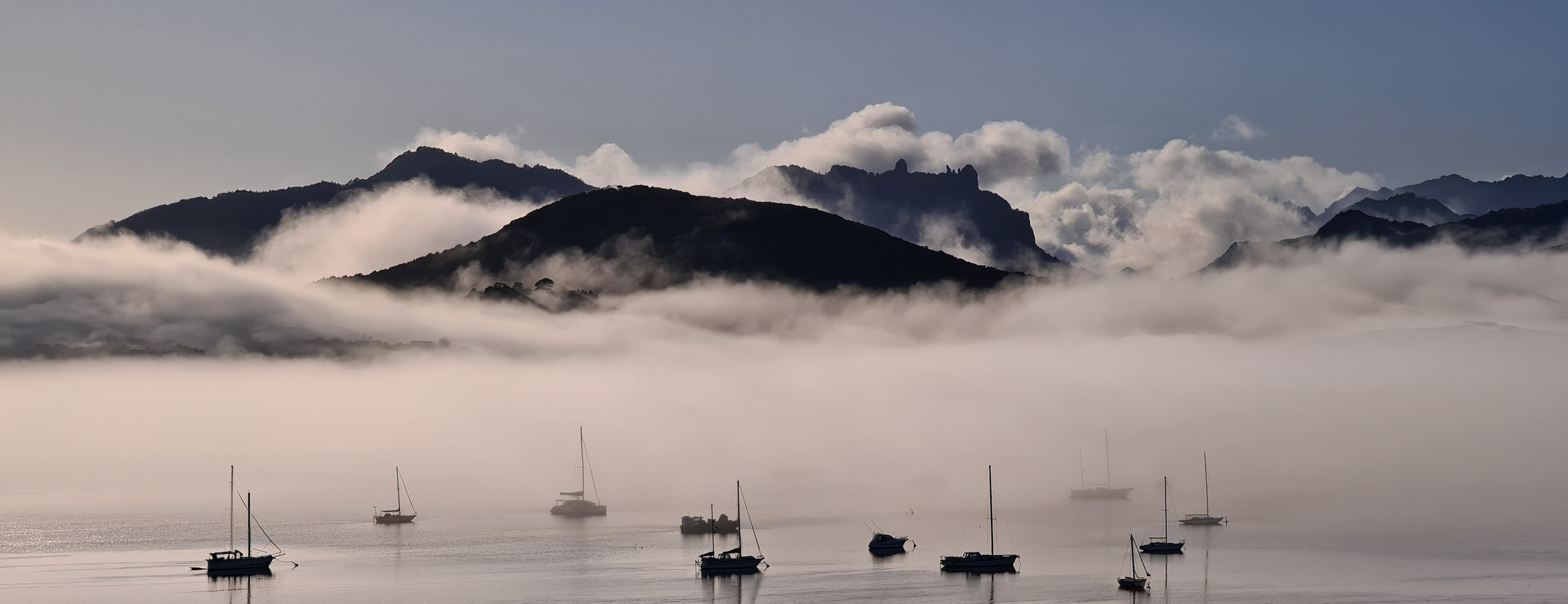 Sailboats anchored in a calm body of water with misty mountains and clouds in the background.