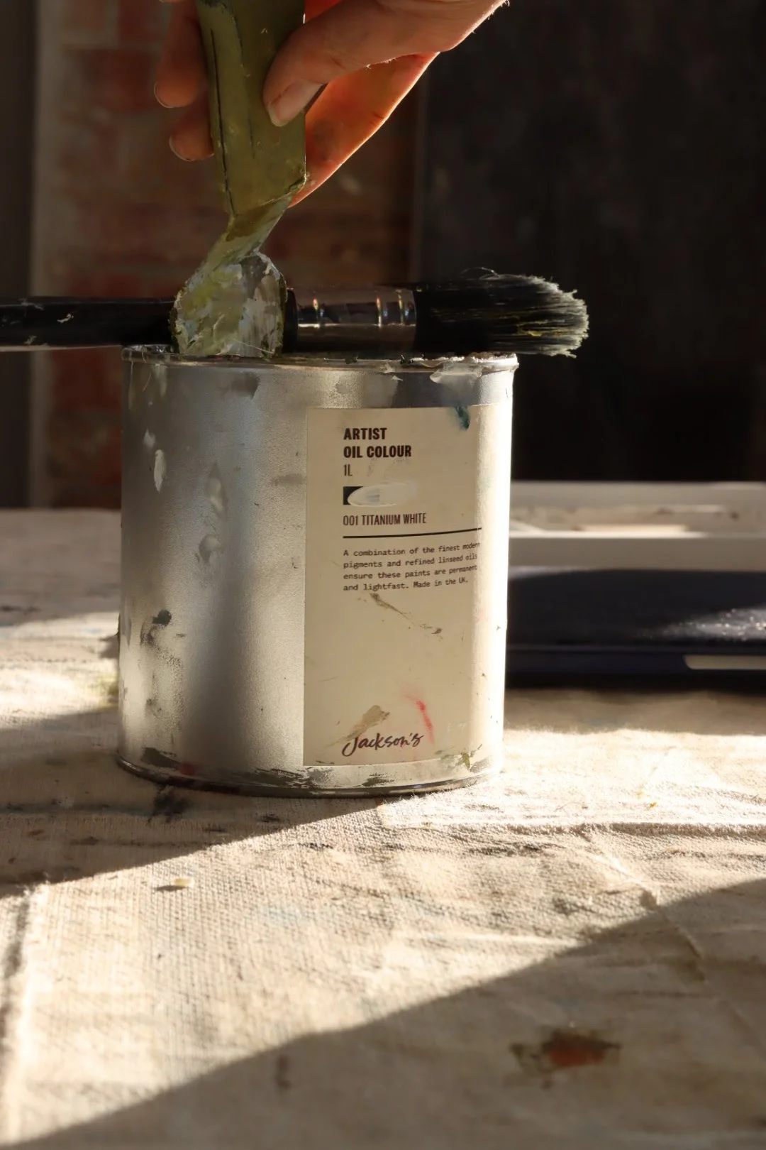 A hand holding a spatula with paint over a can of white paint on a table.