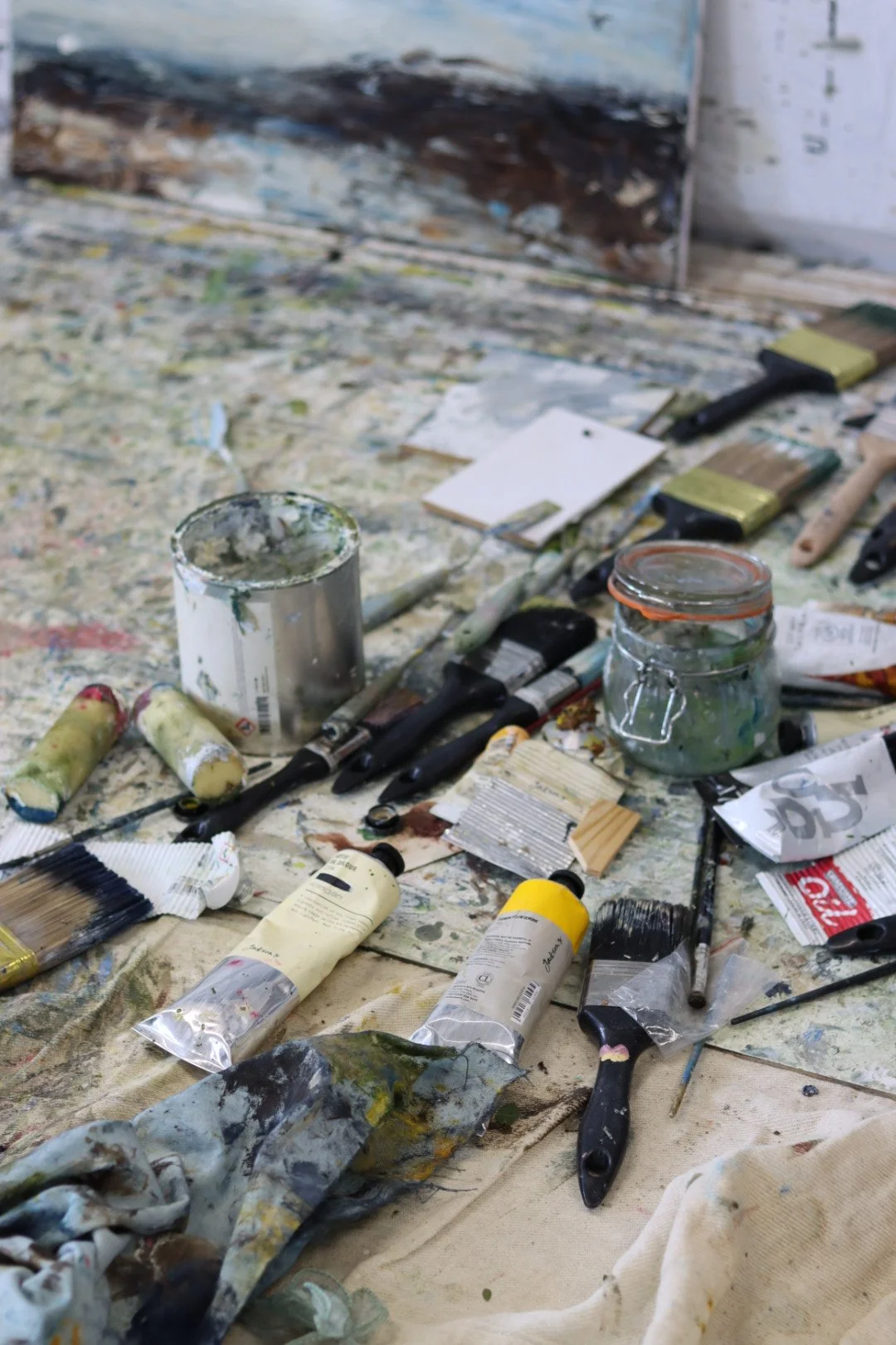 An artist's workspace with various used paint tubes, brushes, and jars on a paint-splattered surface.