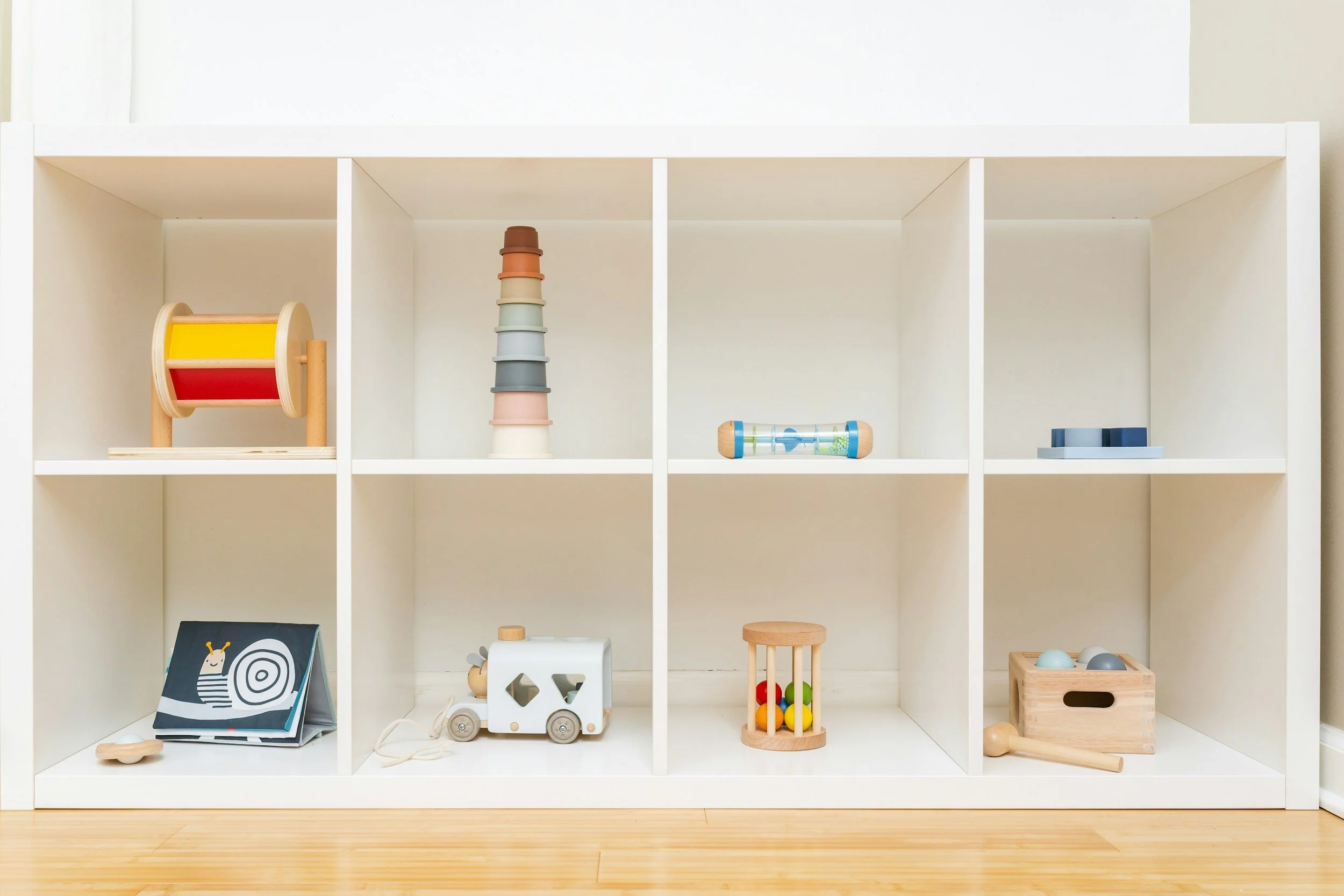 A white cubby shelf with various children's toys and objects, including a color stacker, a toy truck, a wooden rattle, a wire maze with colorful beads, and a framed picture, on a wooden floor.