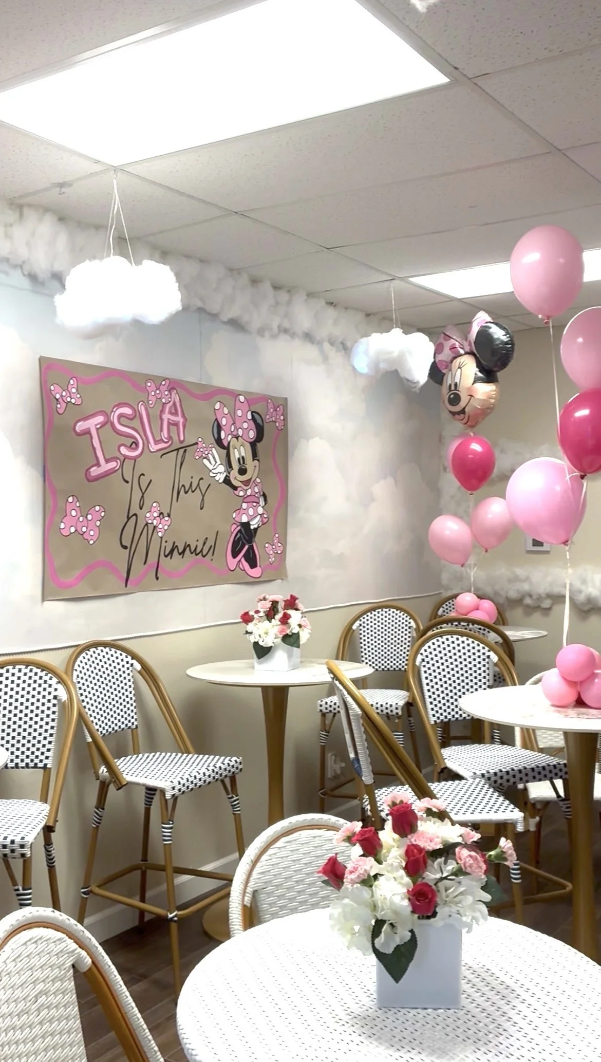 Decorated party room with pink balloons, construction paper Minnie Mouse poster, and flower arrangements on tables.