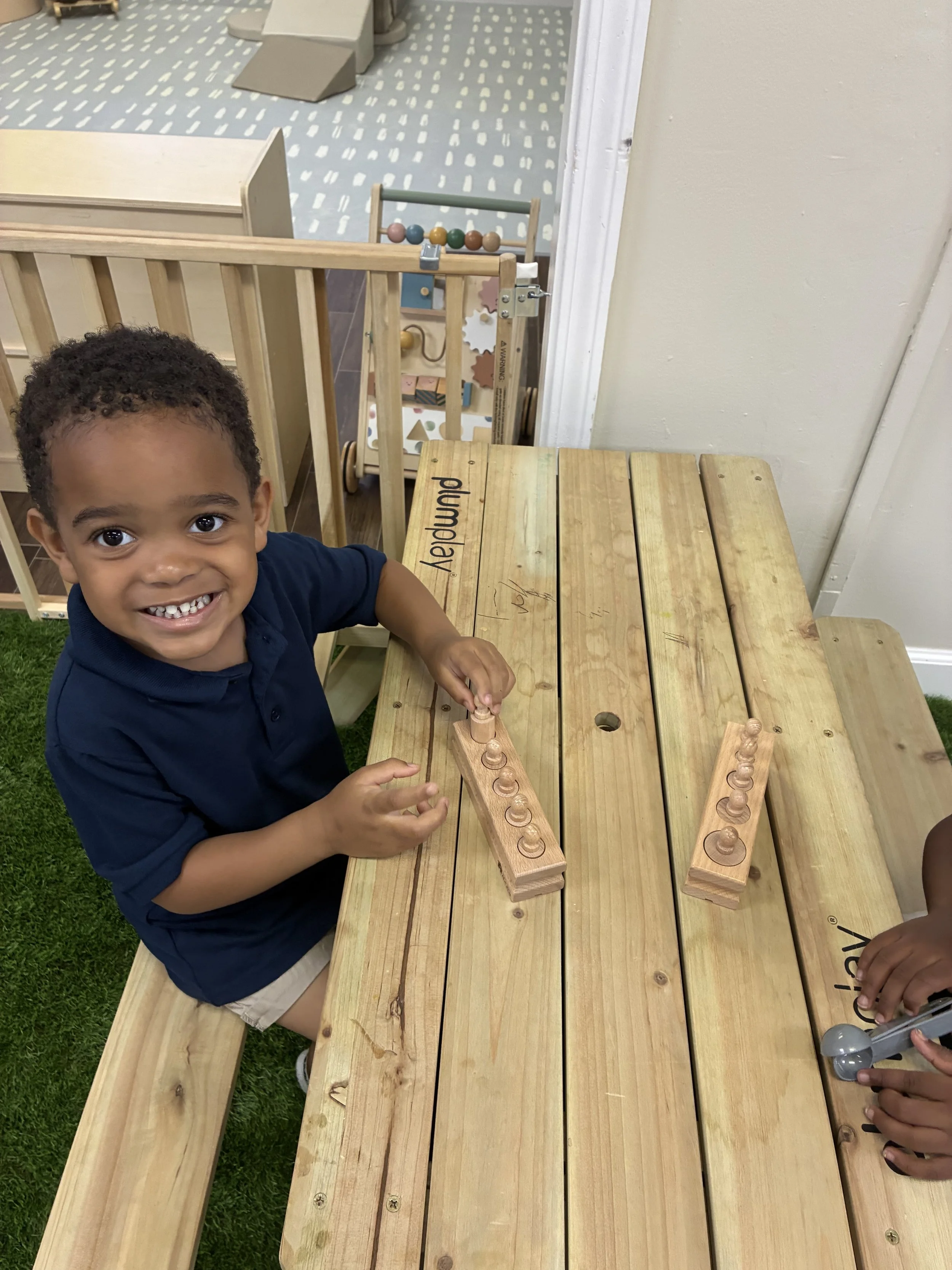 A young boy with curly hair smiling at the camera while playing with wooden stacking pegs in a play area with wooden tables and a toy rack in the background.