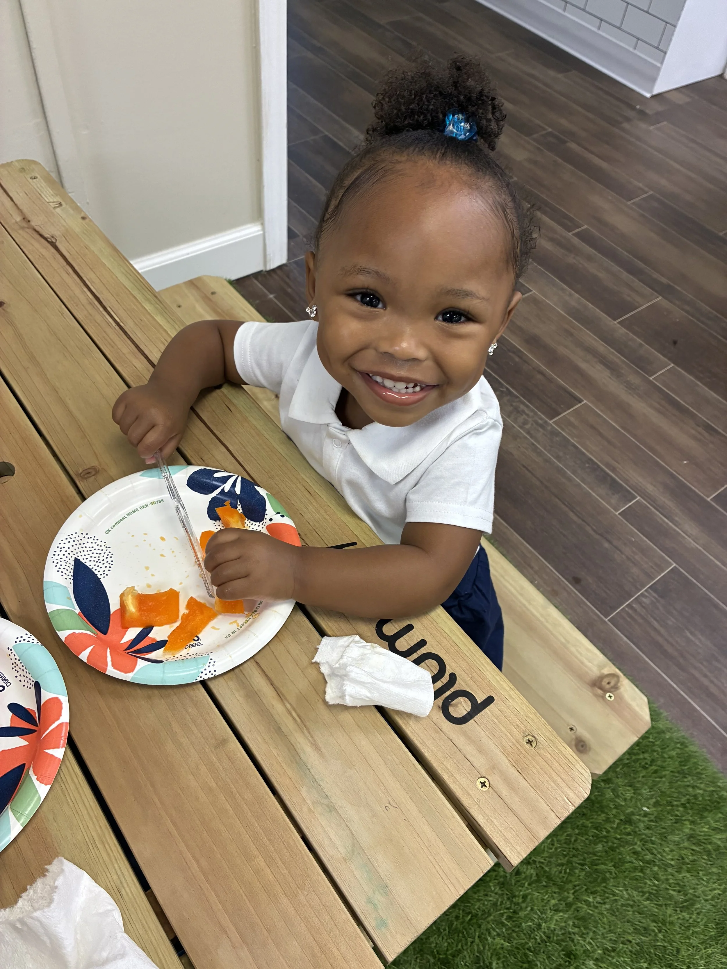 A smiling young girl with curly hair in a ponytail secured with a blue bow, sitting at a wooden table with paper plates and leftover food, looking up at the camera.