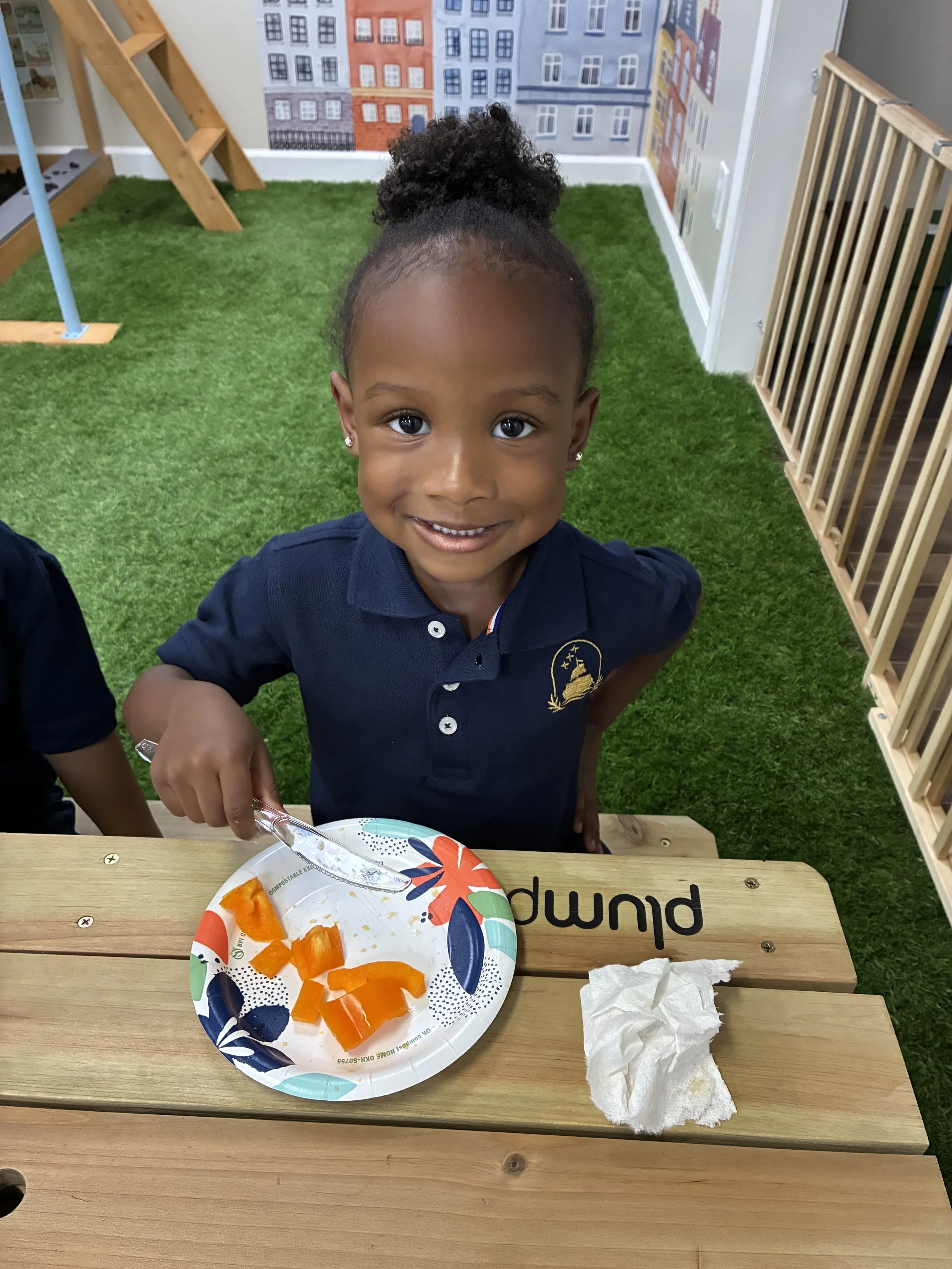A young girl with a happy expression, curly hair styled in a high puff, wearing a navy blue shirt, sitting at a wooden table with a paper plate of sliced orange and yellow bell peppers, in a room with green artificial grass flooring and a colorful mural of city buildings on the wall.