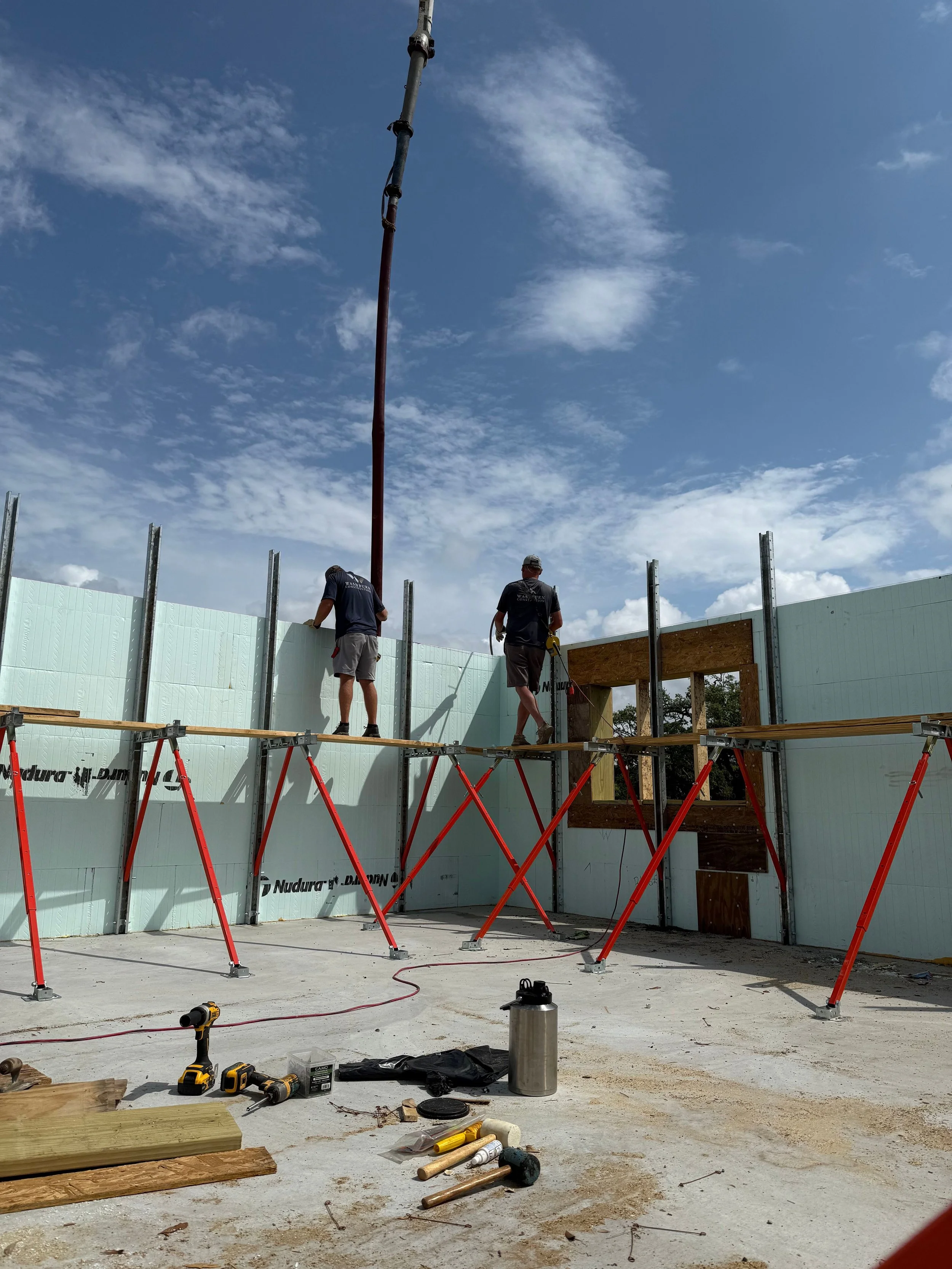 Construction workers building a wall inside a house. Tools and equipment are scattered on the concrete floor, with partial wall framing and insulation visible.